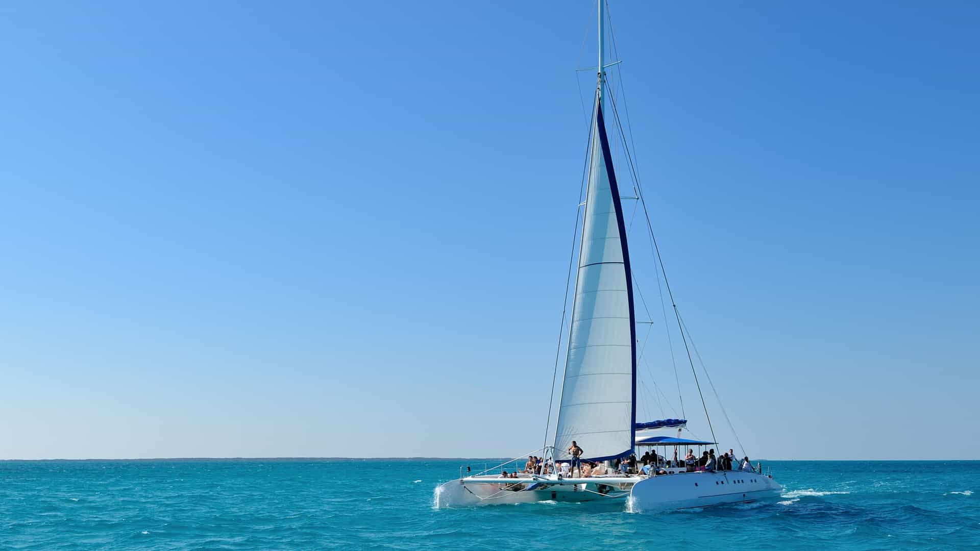 A large white catamaran sailboat glides across the brilliant turquoise waters of the Eastern Caribbean. The boat's tall mast and sails are extended, catching the wind. Several passengers are visible on the deck, enjoying the journey. The water is a stunning shade of blue, and the sky is a clear, cloudless blue, creating a vibrant scene of a typical sailing excursion in the Eastern Caribbean.