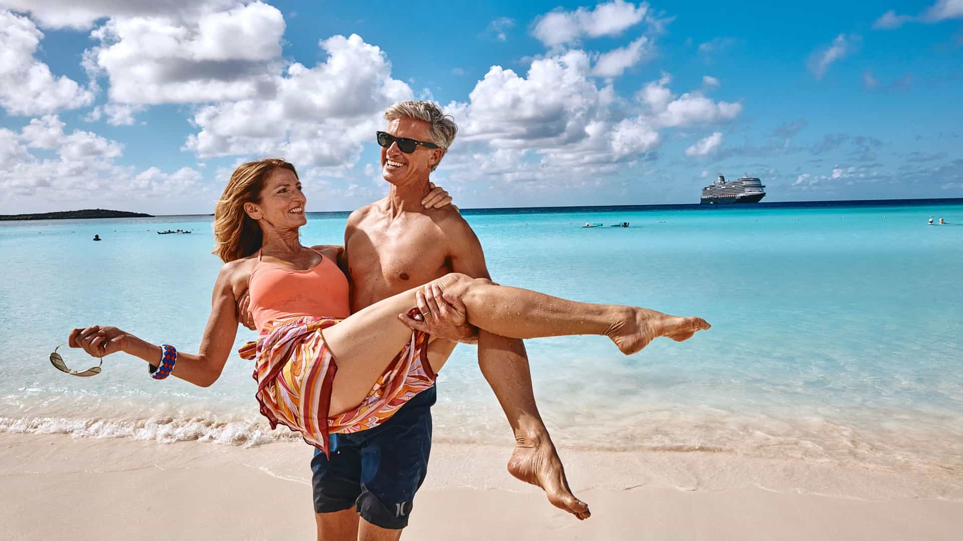 A smiling man carries a woman in his arms on a sunny beach with crystal-clear turquoise water and a cruise ship anchored in the distance.