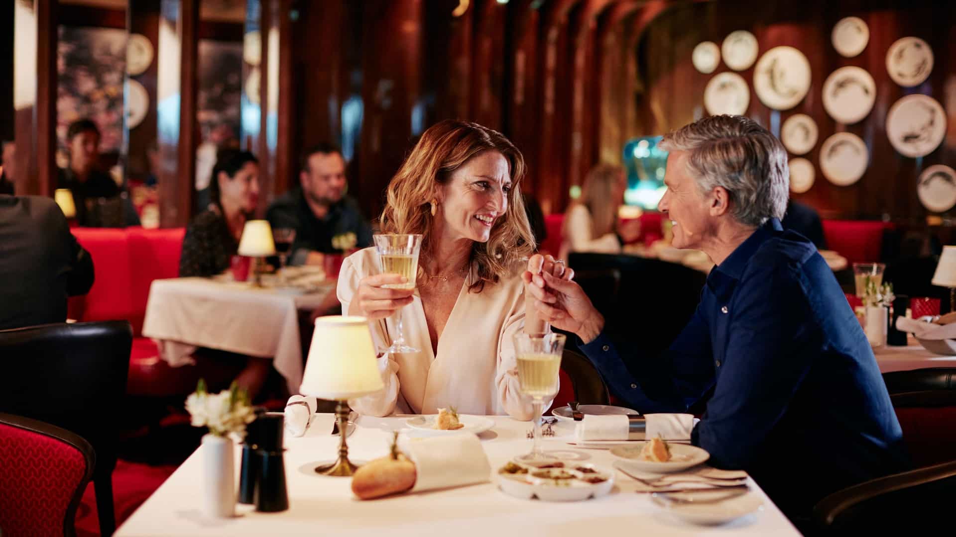 A couple on the Holland America Line's Nieuw Statendam ship is holding hands and looking at each other over dinner and drinks in a restaurant with red booths and chairs.