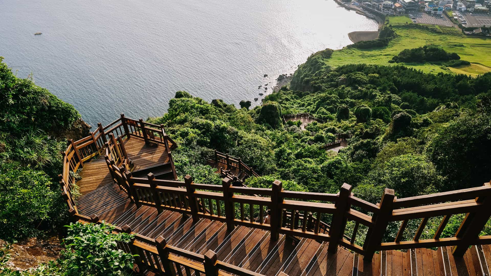 Wooden stairs at Seongsan Ilchulbong on Jeju Island. descending steeply down a lush green cliffside with a wooden railing, offering a panoramic view of the sparkling blue ocean and a coastal town in the distance.