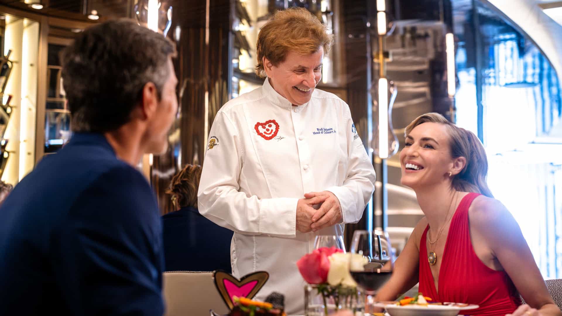 Chef Rudi Sodamin in a white chef's coat smiles and chats with a couple enjoying a meal at a table in a specialty restaurant.