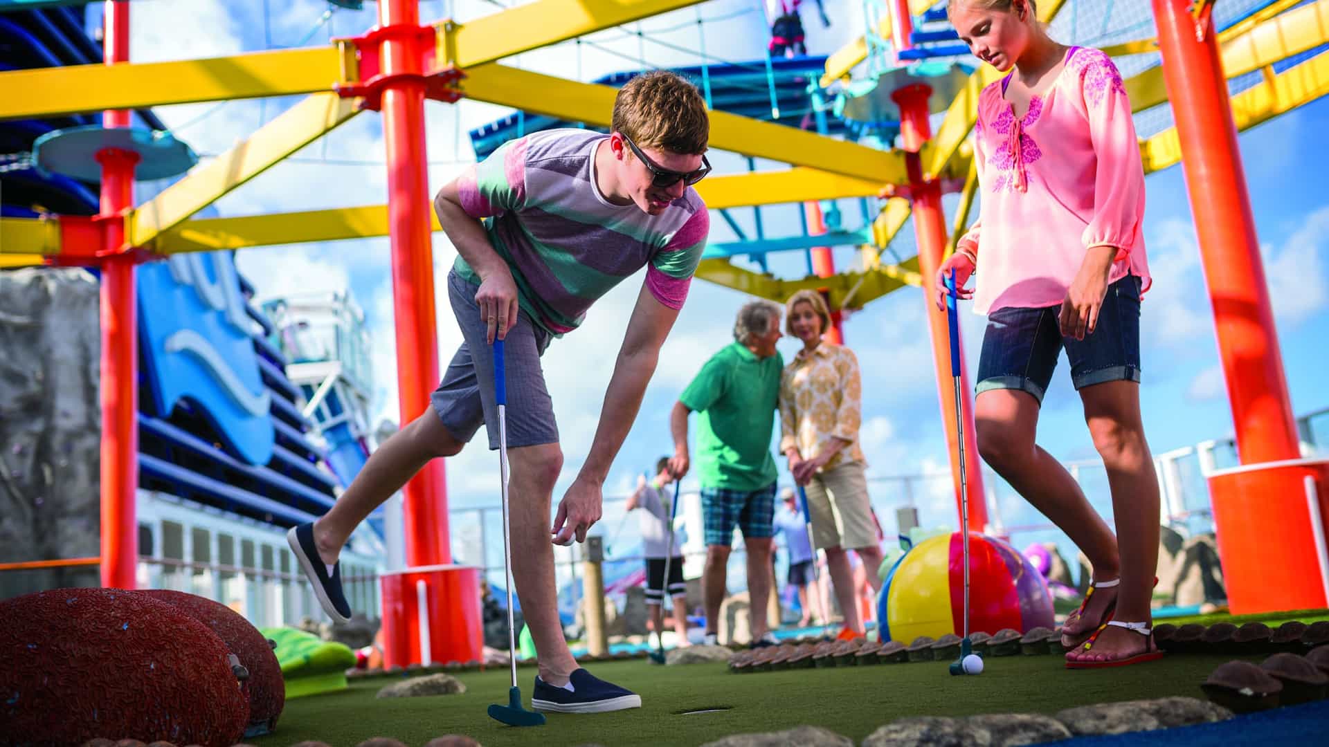 Two teens playing mini golf on the top deck of Norwegian Breakaway, with the ship's colorful ropes course structure visible overhead — one of the complimentary onboard activities available to families on Norwegian Cruise Line.