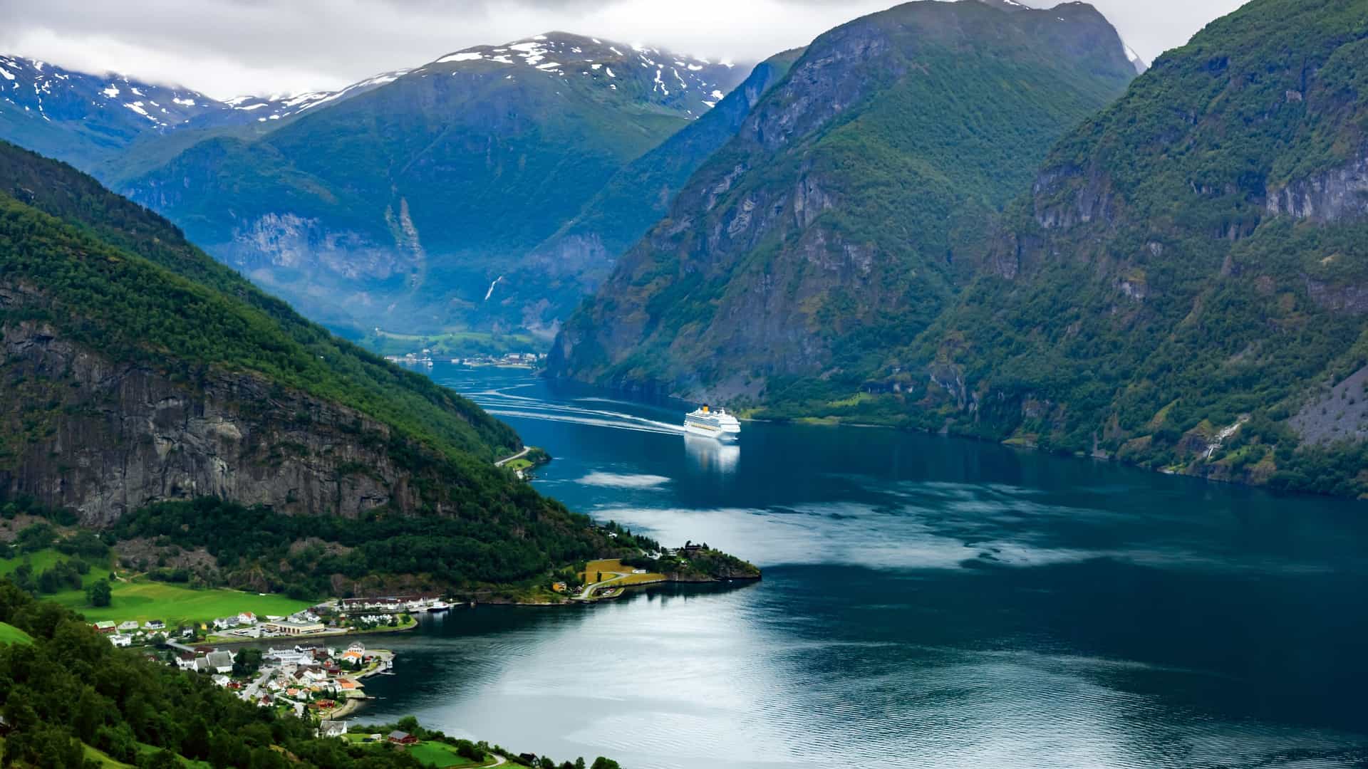 A panoramic, high-angle view of the Geirangerfjord in Norway. A white cruise ship navigates the dark blue water, passing a small, clustered village with green fields on the near shore. Towering, steep mountains covered in lush green forest flank the fjord, with snow-capped peaks visible in the background under a cloudy sky.