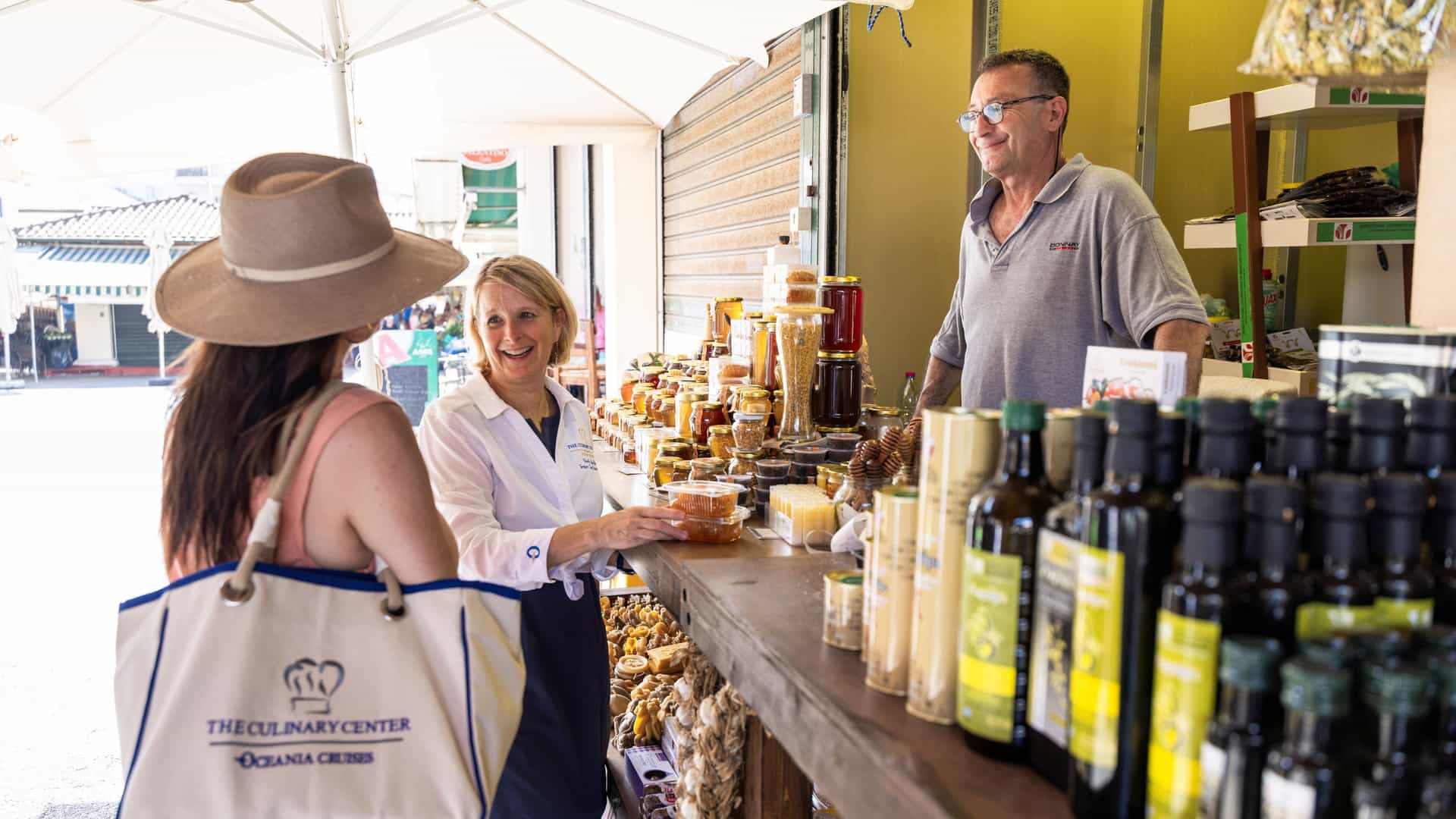 An Oceania Cruises Culinary Center chef instructor and guest browsing local honey, olive oil, and specialty products at an outdoor market stall during a Culina…An Oceania Cruises Culinary Center chef instructor and guest browsing local honey, olive oil, and specialty products at an outdoor market stall during a Culinary Discovery Tour in Corfu, Greece.