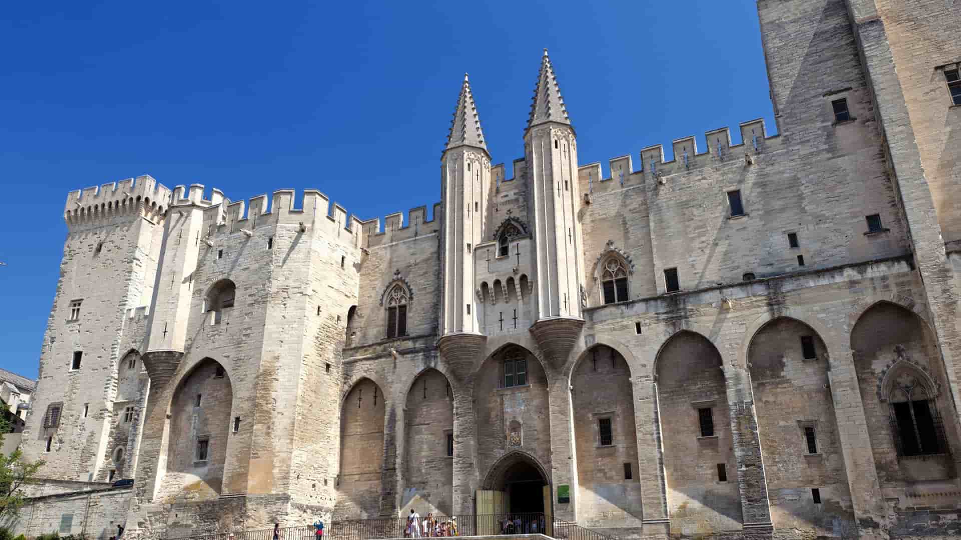 Palace of the Popes, Avignon, France: historic stone walls, arched entrances, twin spires under blue sky.
