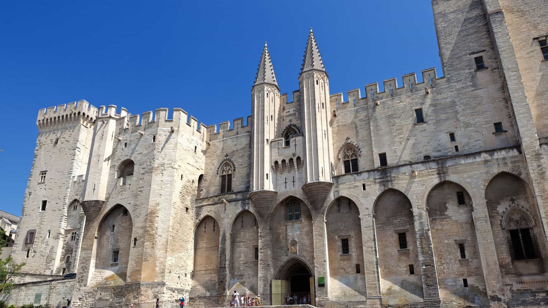 Palace of the Popes, Avignon, France: historic stone walls, arched entrances, twin spires under blue sky.