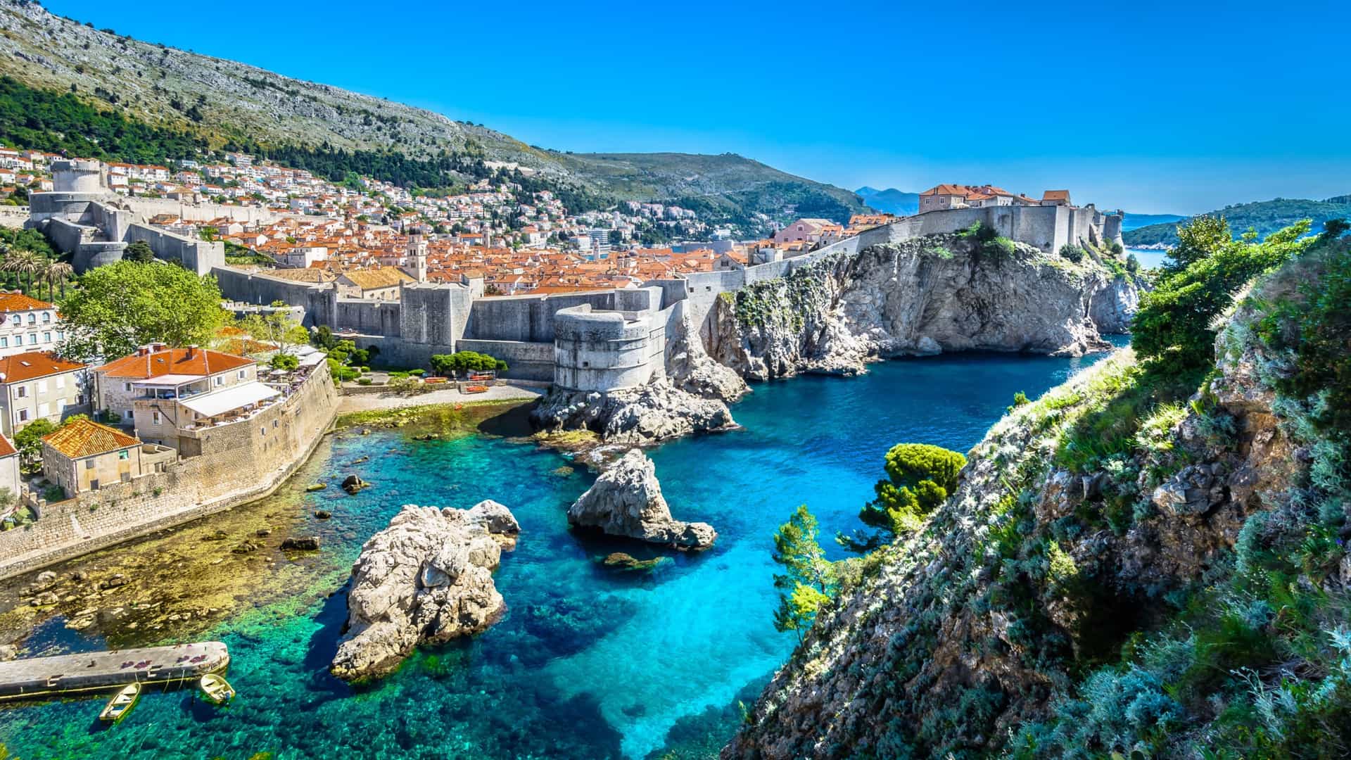 A scenic view of Dubrovnik's Old Town, with its medieval city walls and terracotta roofs, perched on cliffs overlooking the clear turquoise waters of the Adriatic Sea.