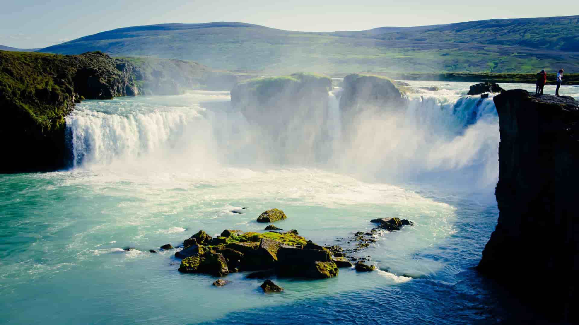 A breathtaking view of Goưafoss, the "Waterfall of the Gods," in northern Iceland, with powerful cascades of turquoise water and two people standing on the rocky cliff.
