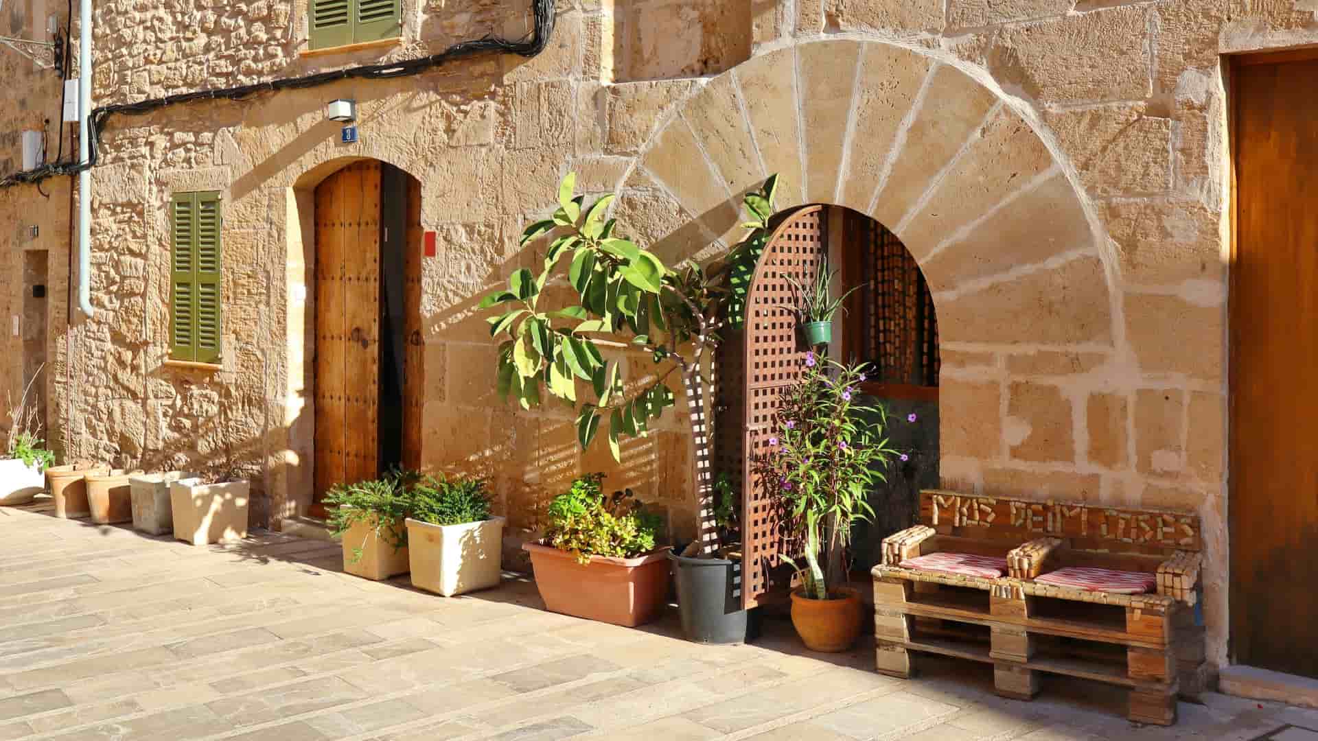 A sunlit, narrow street scene in Alcudia, Mallorca, with an arched stone doorway, potted plants, and a rustic wooden bench.