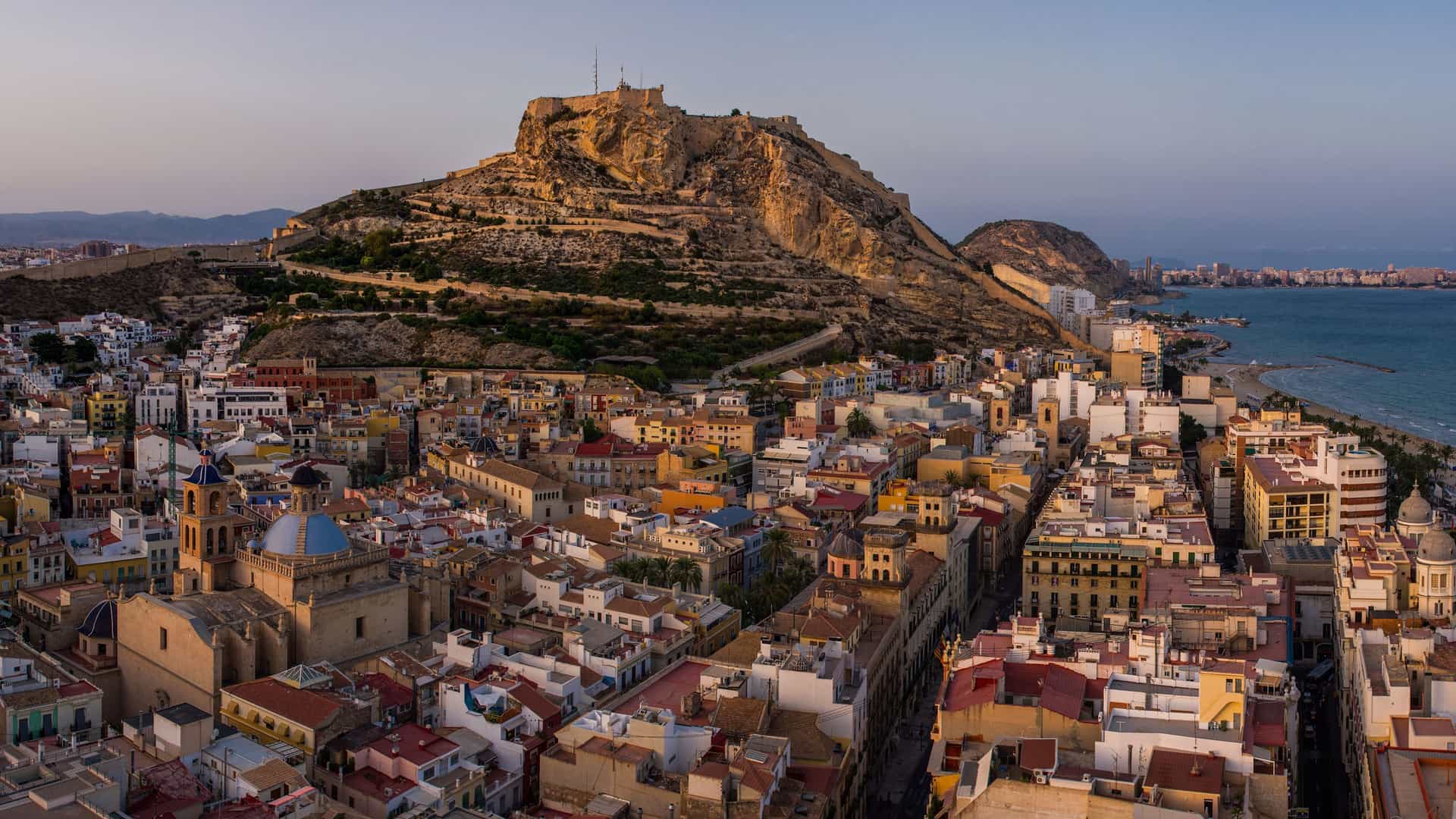 A stunning panoramic view of Alicante, Spain, at sunset, with the historic city nestled at the foot of Mount Benacantil and the Santa Bárbara Castle, with the Mediterranean Sea in the distance.