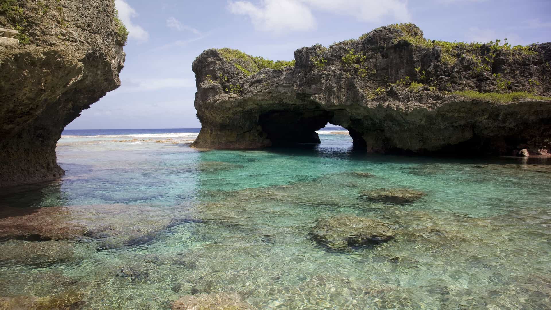 A stunning natural rock pool with an archway carved into the rock, leading to the open ocean, on the island of Alofi. The water is clear and shallow, with a rocky bottom visible beneath the surface.