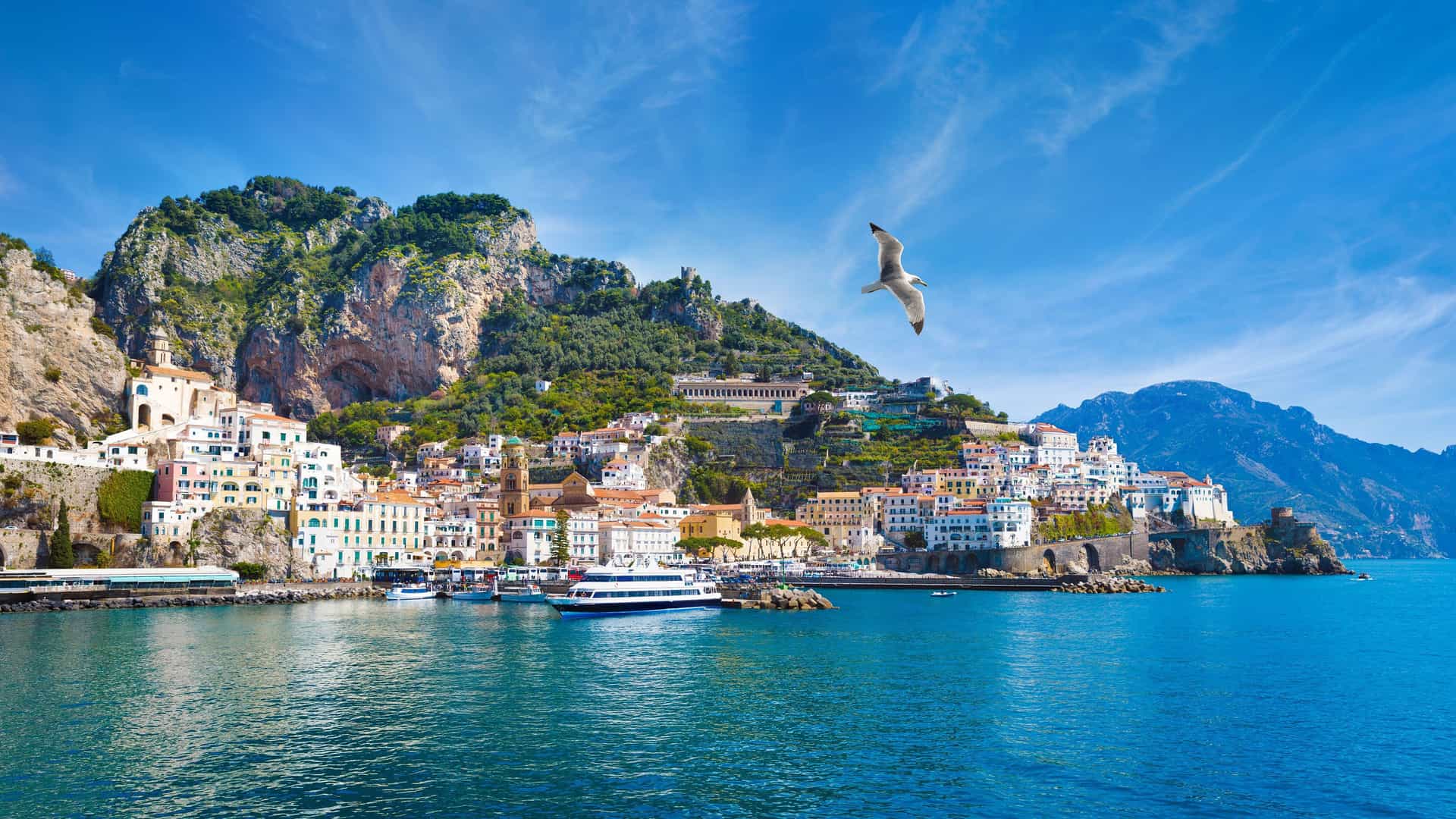 A stunning panoramic shot of the Amalfi Coast's colorful, terraced houses built into the dramatic cliffs overlooking the turquoise Mediterranean Sea, with a seagull flying overhead.