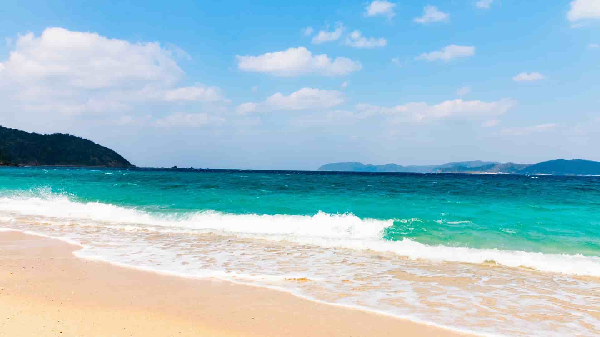 A sunny seascape of a sandy beach in Amami Ōshima, Japan, with turquoise waves crashing onto the shore and a lush, green island in the distance.