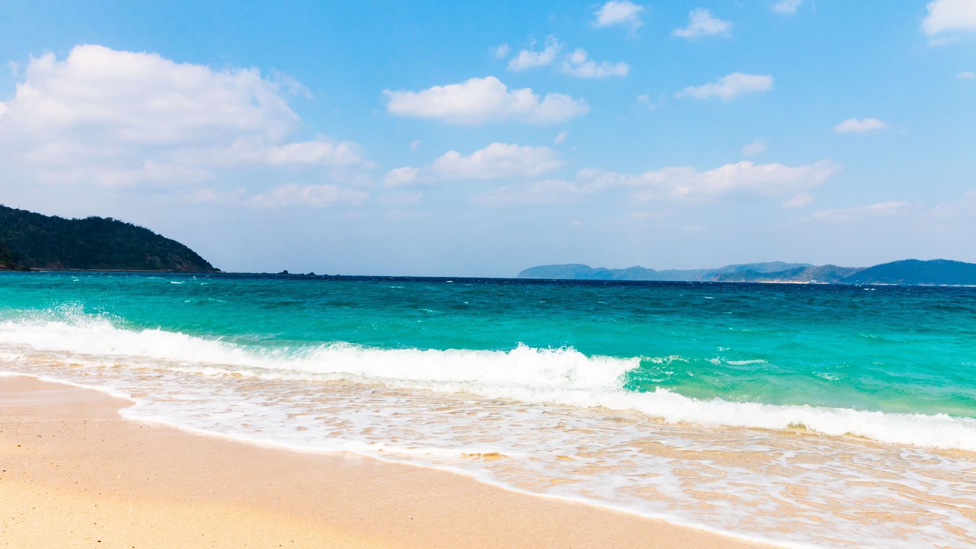 A sunny seascape of a sandy beach in Amami Ōshima, Japan, with turquoise waves crashing onto the shore and a lush, green island in the distance.
