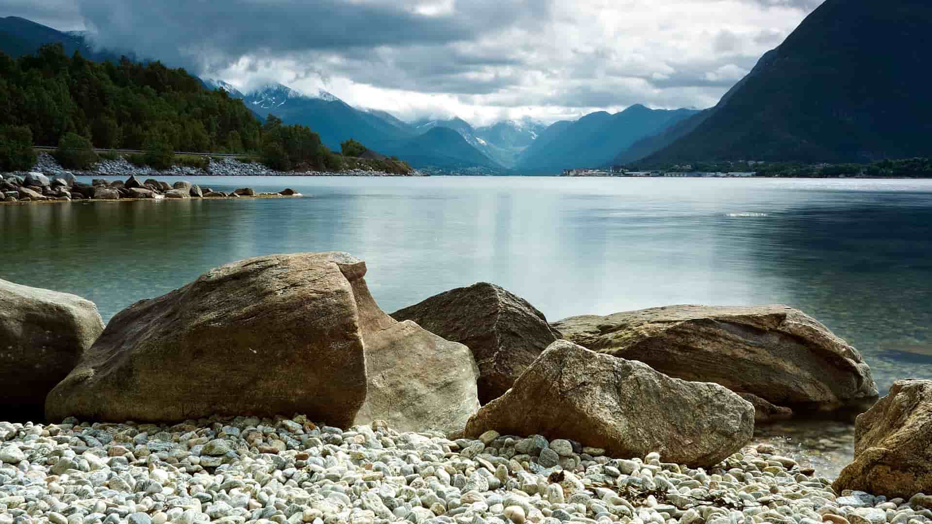 A dramatic landscape of the Romsdalsfjord in Ć
ndalsnes, Norway, with large rocks in the foreground and majestic snow-capped mountains and a cloudy sky in the background.