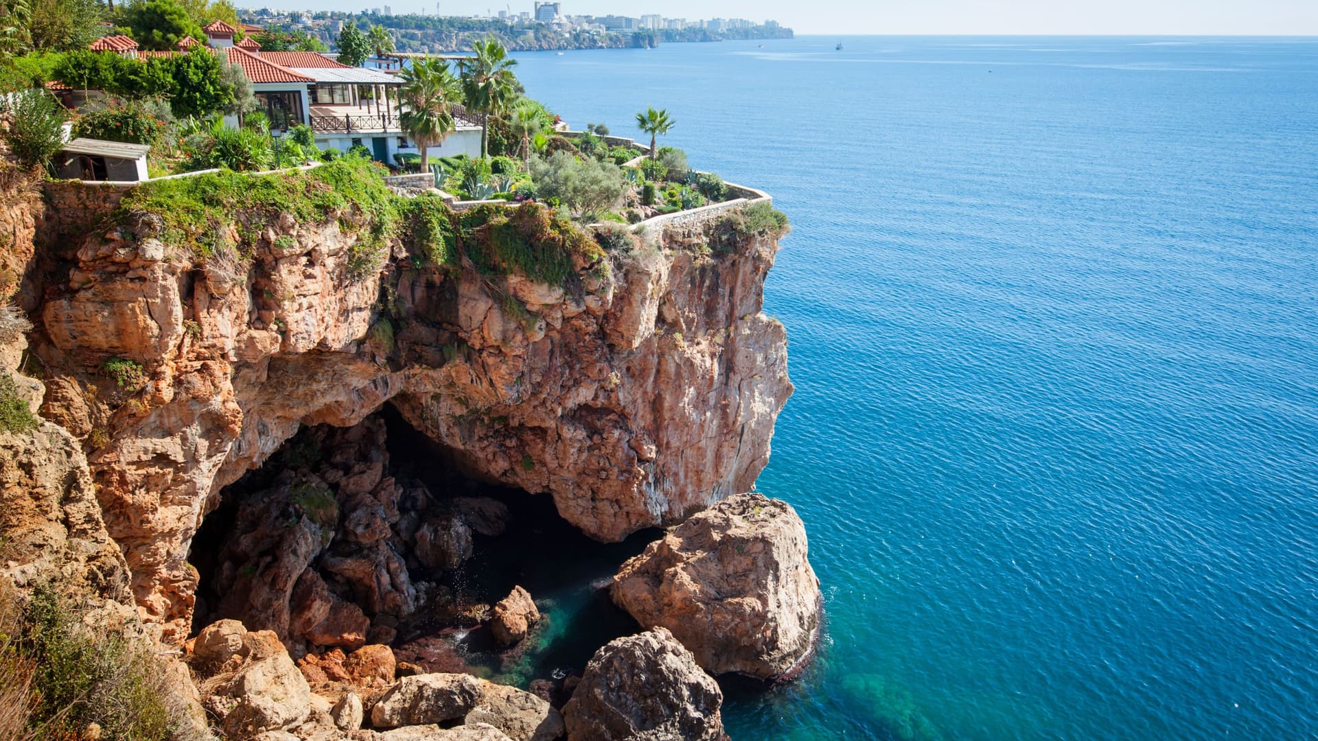 A sunny cliffside view of the clear blue Mediterranean Sea in Antalya, Turkey, with a beautiful house and palm trees perched on the edge of the rocky cliffs.