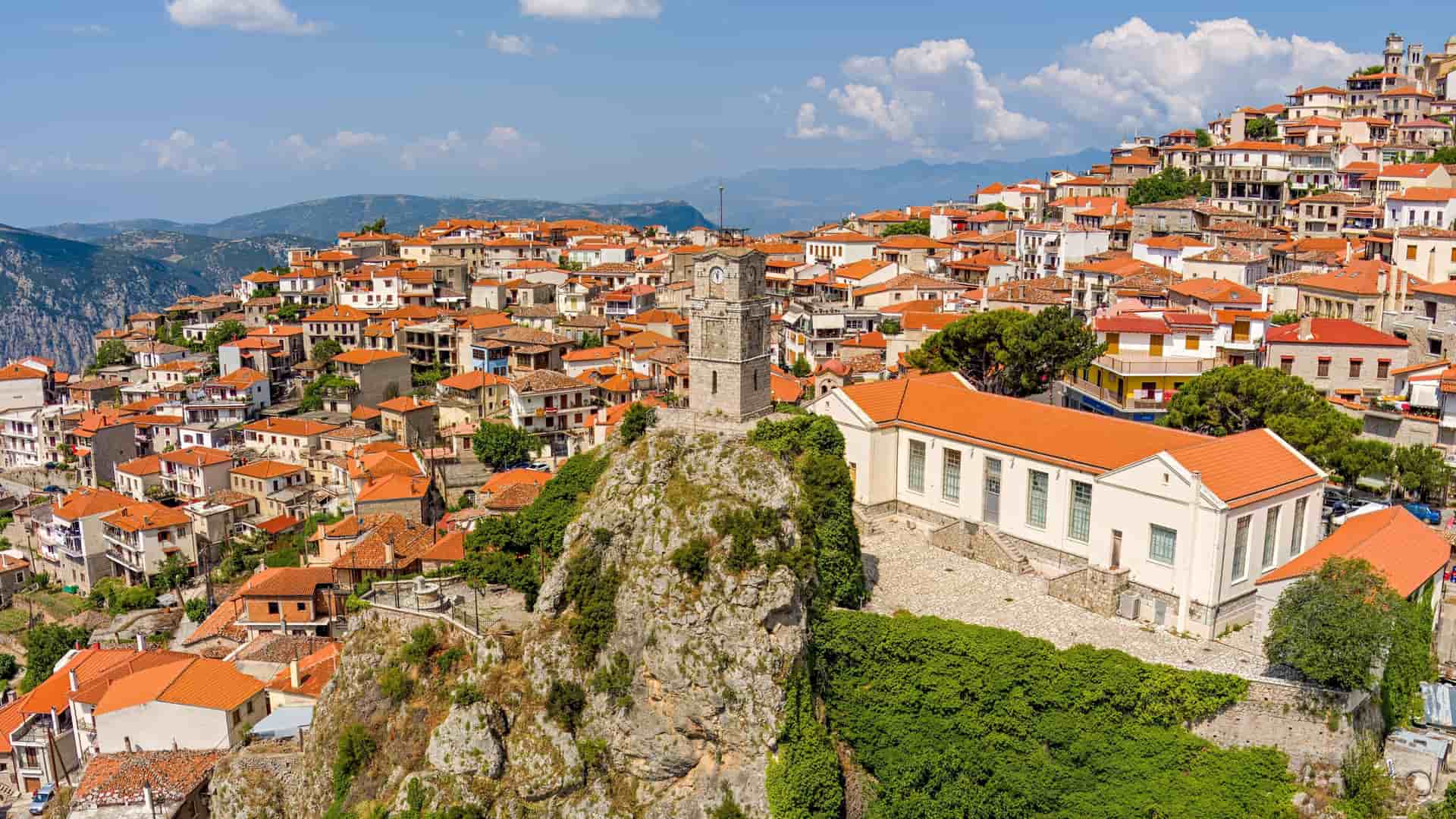 An aerial view of Arachova, Greece, showing the scenic mountain town built on a slope, with the iconic Clock Tower rising from a rocky outcrop.