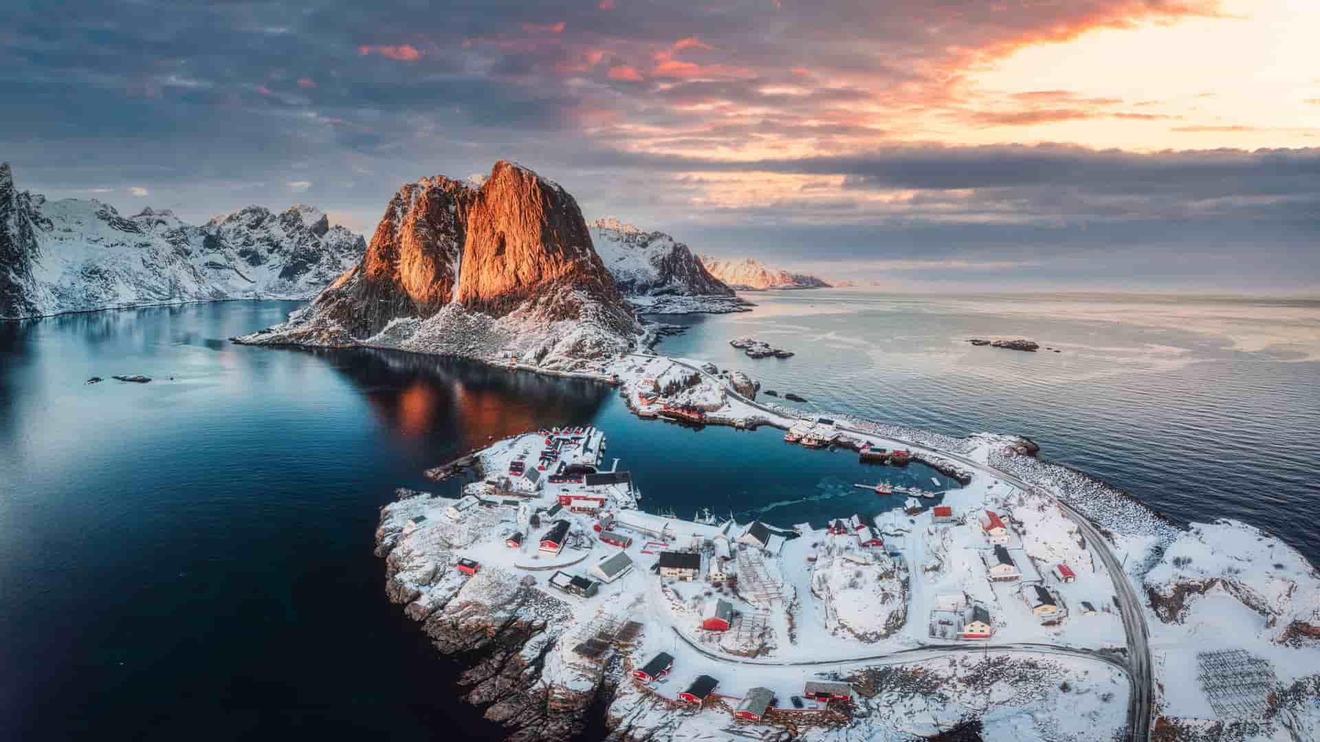 An aerial panoramic view of a snow-covered fishing village on the Arctic Circle, with a massive mountain peak rising from the sea at sunset.