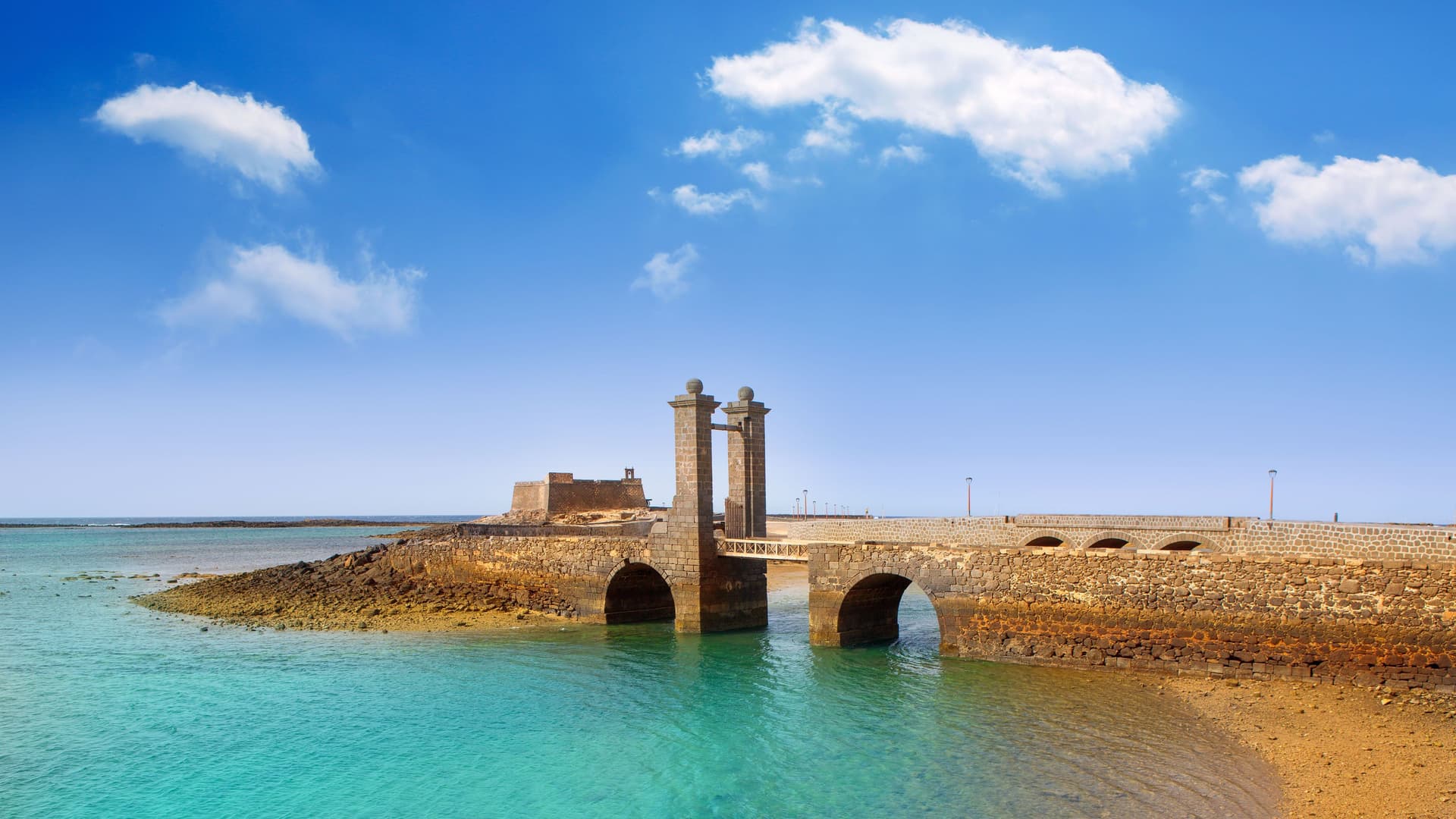 A scenic view of the Puente de las Bolas, a historic stone bridge with two decorative spheres, leading to the Castillo de San Gabriel fortress in Arrecife, Lanzarote, Canary Islands.