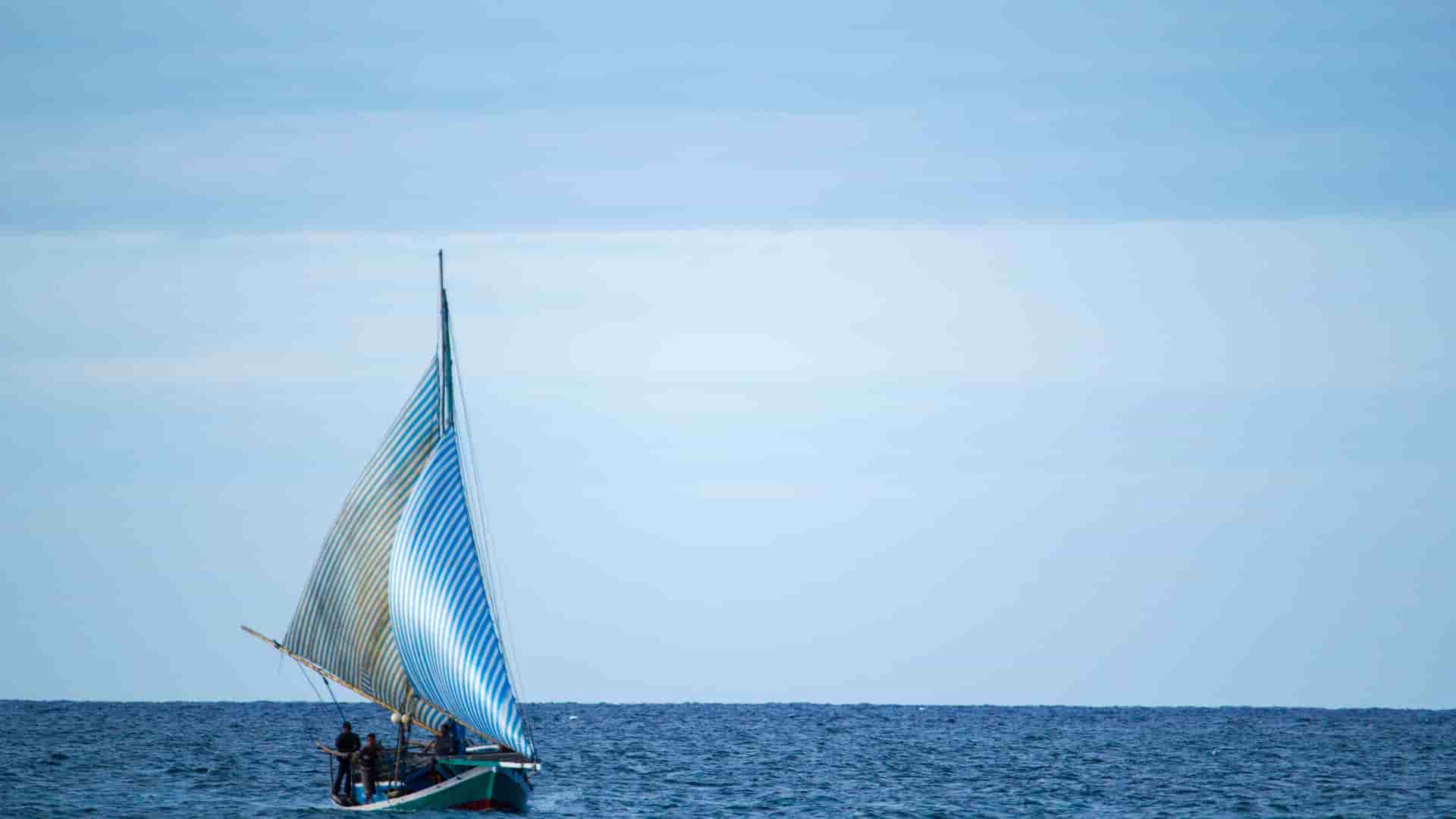 A sailboat with blue and white striped sails sailing on the calm blue waters of the Indian Ocean near Ashmore Reef, Australia, on a hazy day.