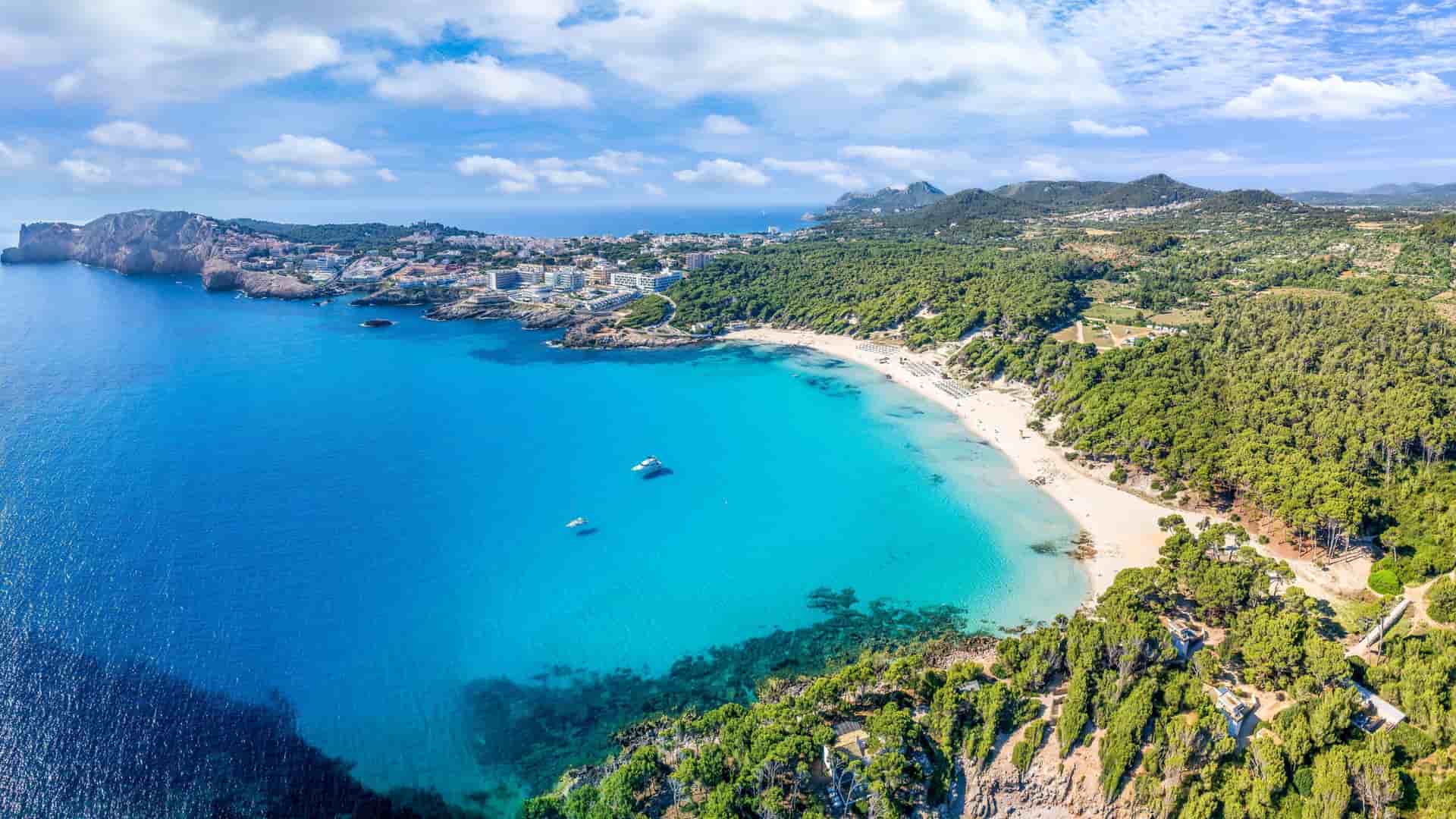 An aerial view of a picturesque beach on the Balearic Sea, with turquoise water and white sand framed by lush green trees and rocky cliffs.