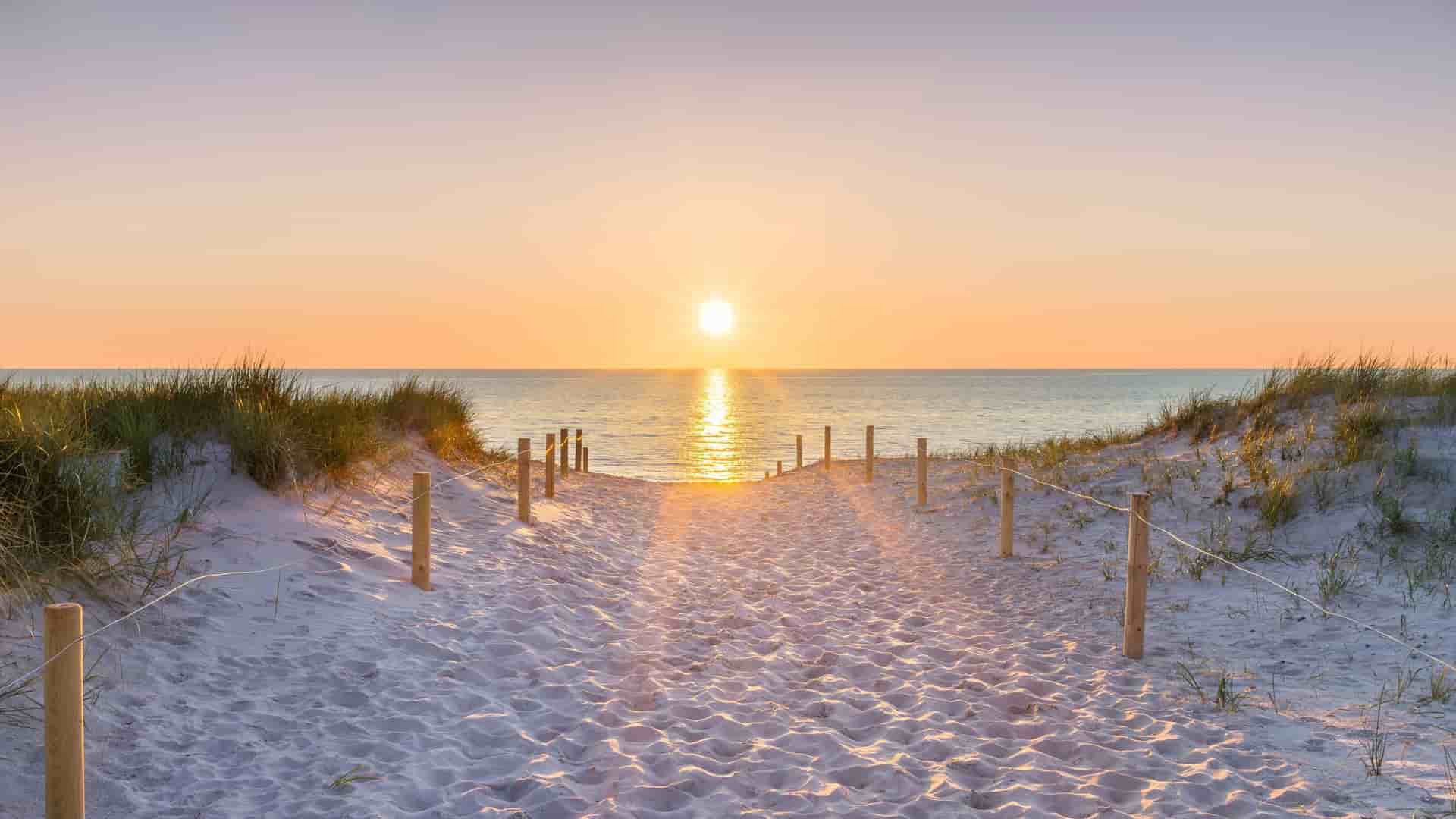A sandy pathway with wooden posts and a rope fence leads over grassy dunes to the Baltic Sea as the sun sets over the horizon.