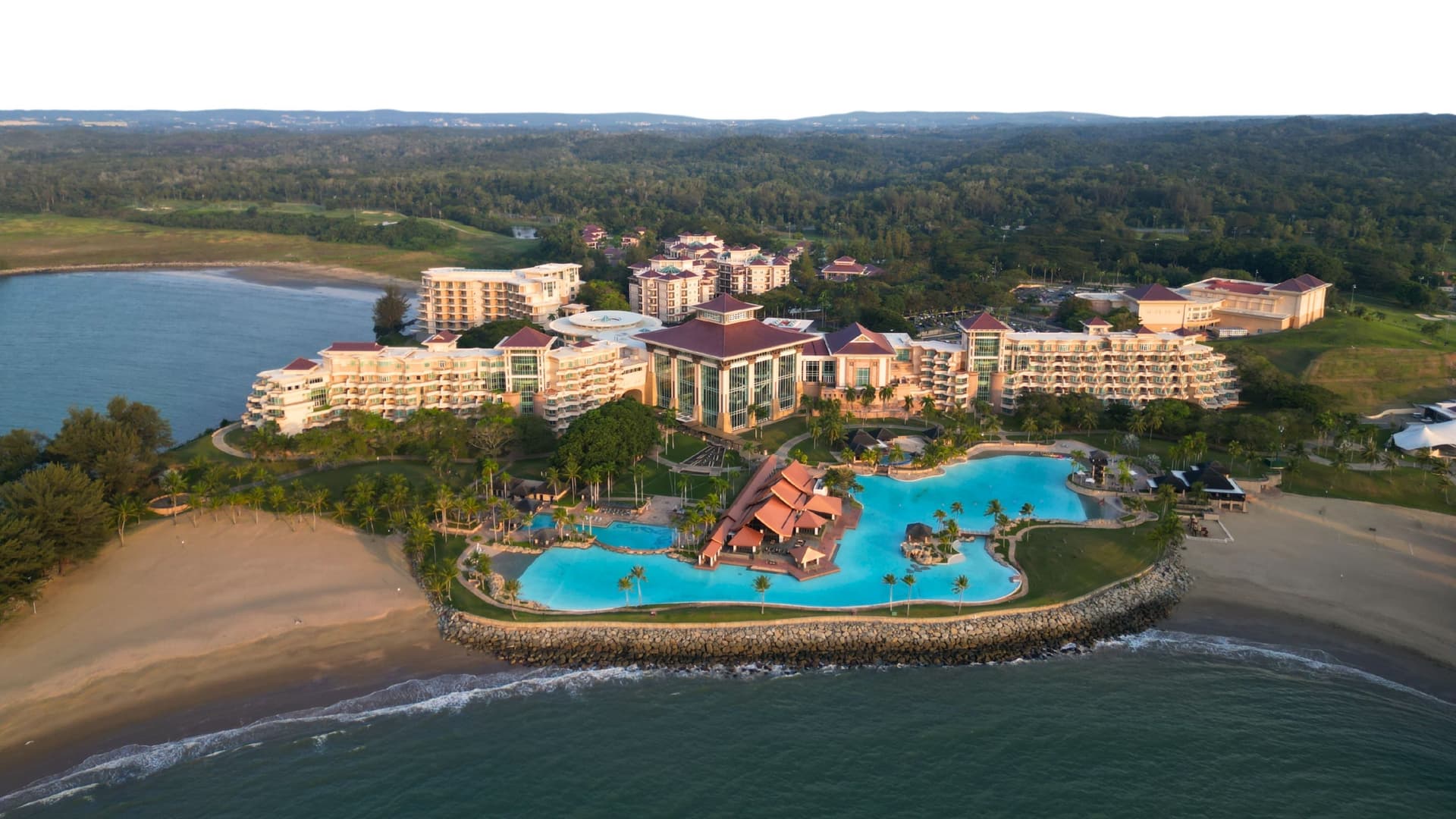 An aerial view of a luxurious resort complex in Bandar Seri Begawan, Muara, with multiple buildings, a large swimming pool, and a private beach.