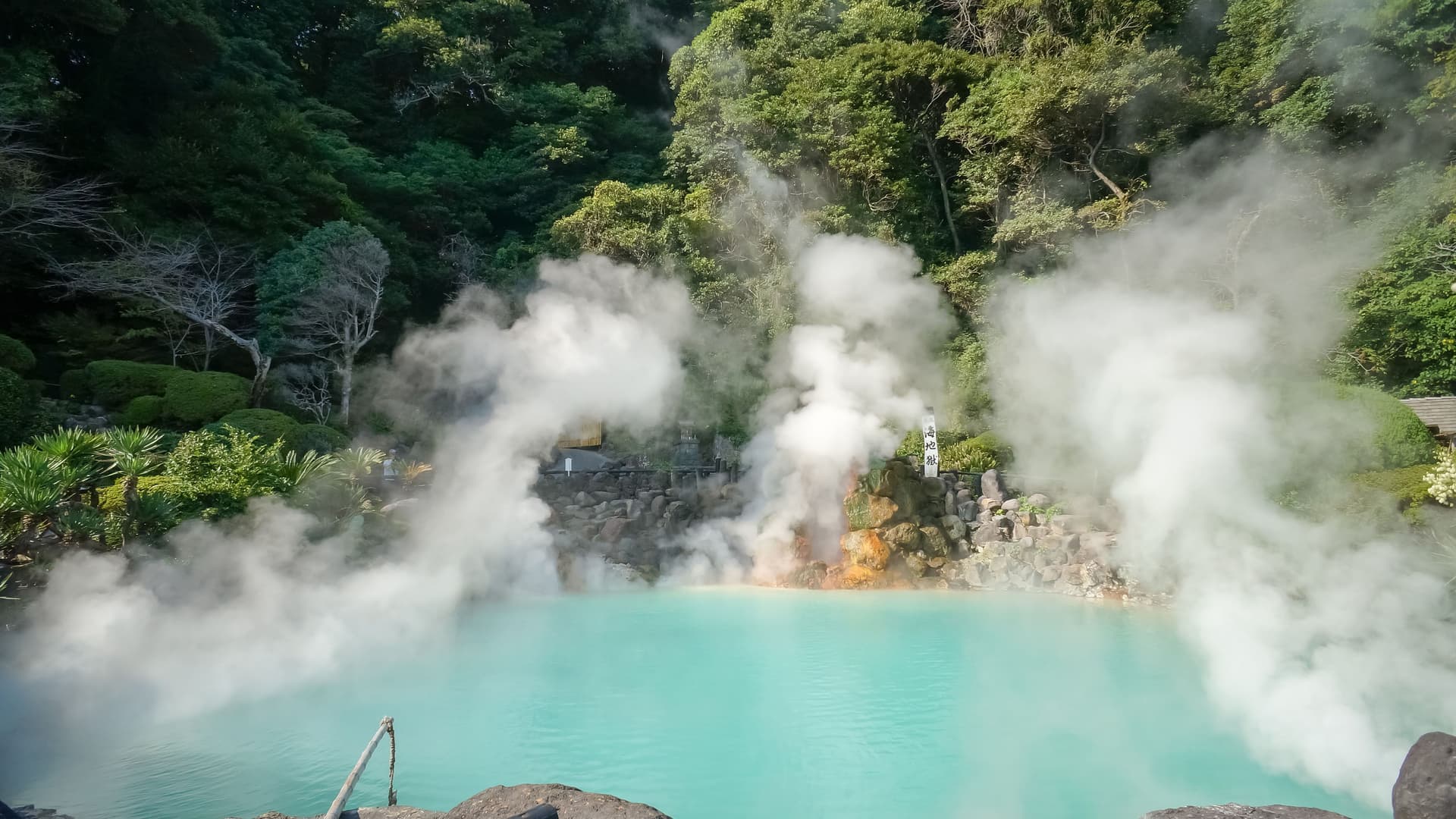 A stunning view of the Umi Jigoku ("Sea Hell") in Beppu, Japan, featuring a vibrant turquoise-blue hot spring lake emitting thick plumes of white steam, surrounded by lush green foliage.