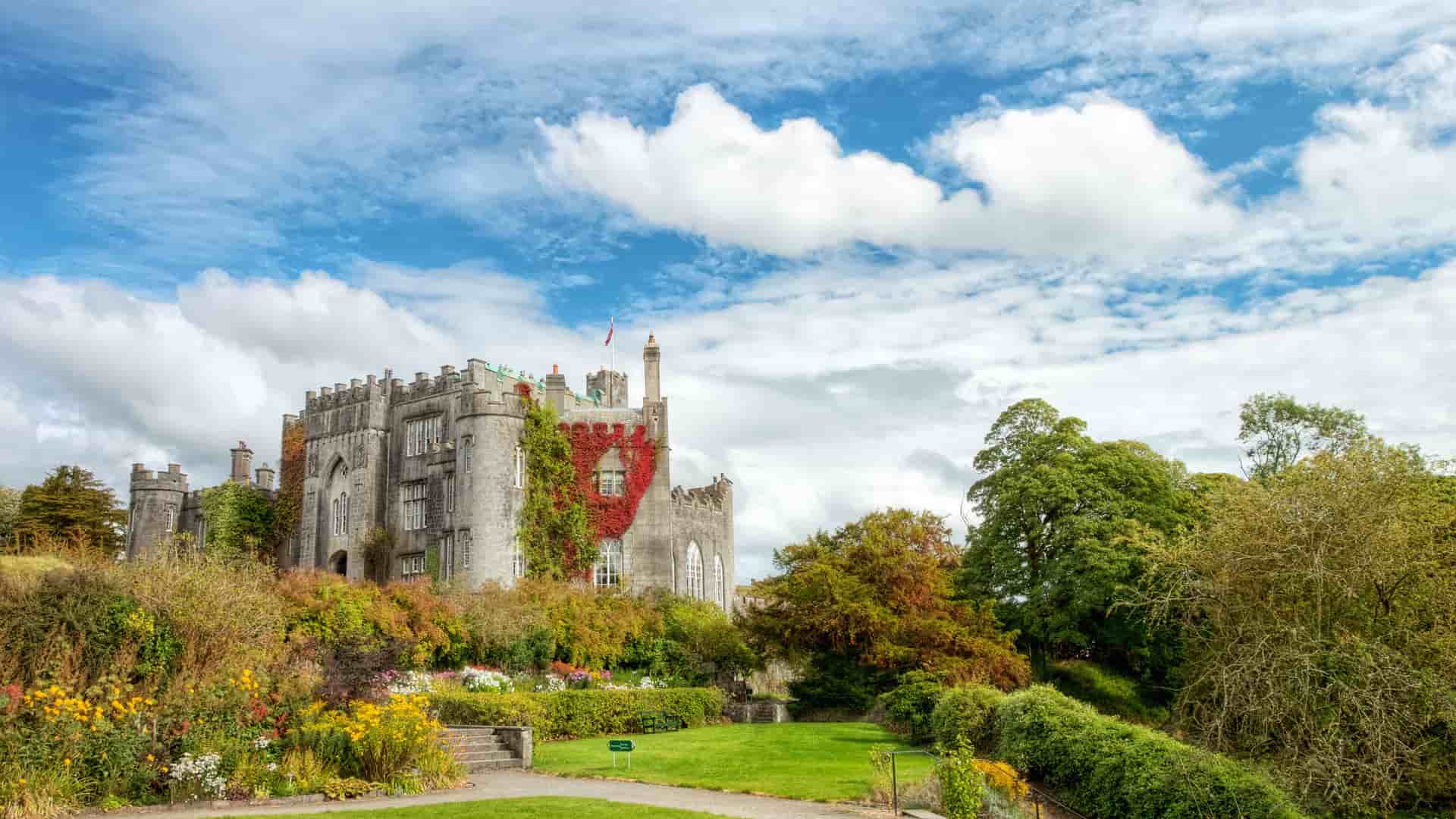 A picturesque view of Birr Castle in Birr, Ireland, a large gray stone building with turrets, partially covered in vibrant red climbing vines. The castle is situated on a grassy hill overlooking a beautiful garden filled with colorful flowers, all under a bright blue sky with large white clouds.