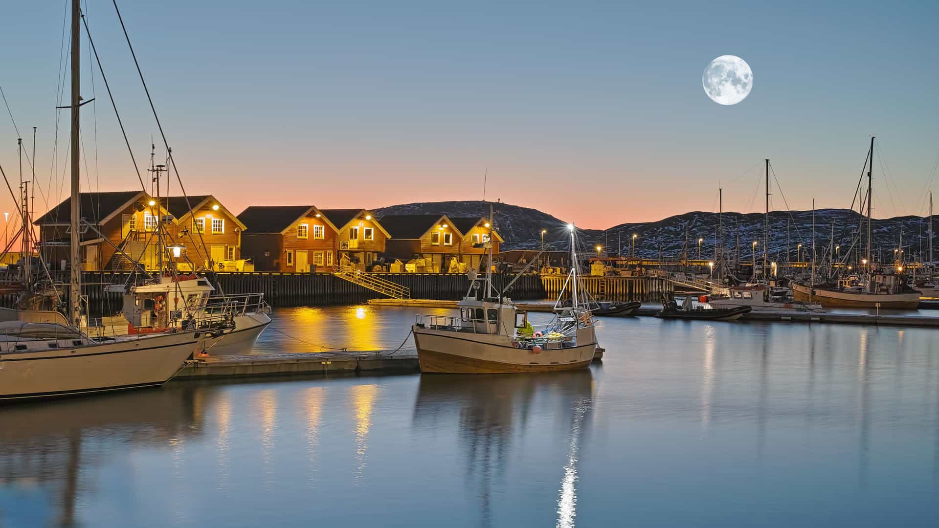 A scenic nighttime view of the harbor in Bodø, Norway, with boats and illuminated waterfront buildings under a full moon and a vibrant twilight sky.