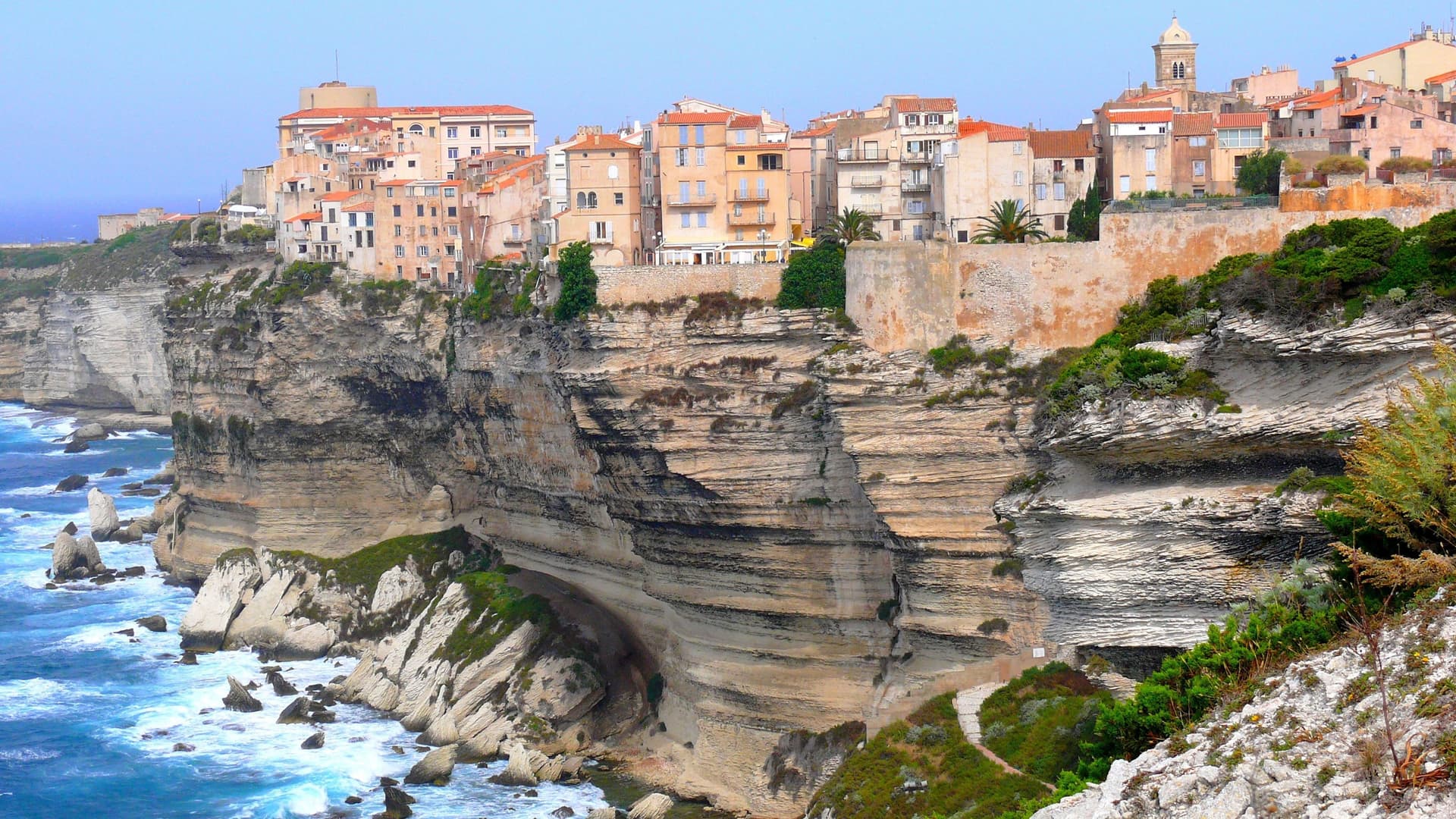 A scenic view of the city of Bonifacio, Corsica, with its historic buildings perched precariously on the edge of a dramatic white limestone cliff overlooking the turquoise Mediterranean Sea.
