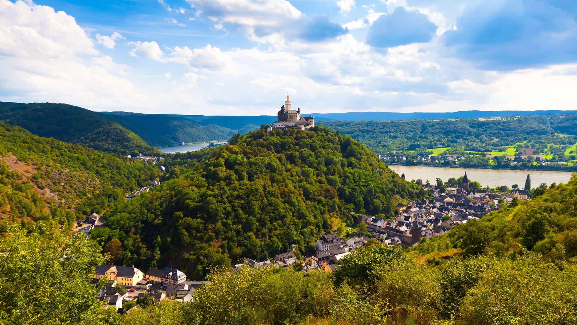 A stunning aerial landscape of Braubach, Germany, with the historic Marksburg Castle majestically situated atop a green, forested hill overlooking the Rhine River and the picturesque town below.