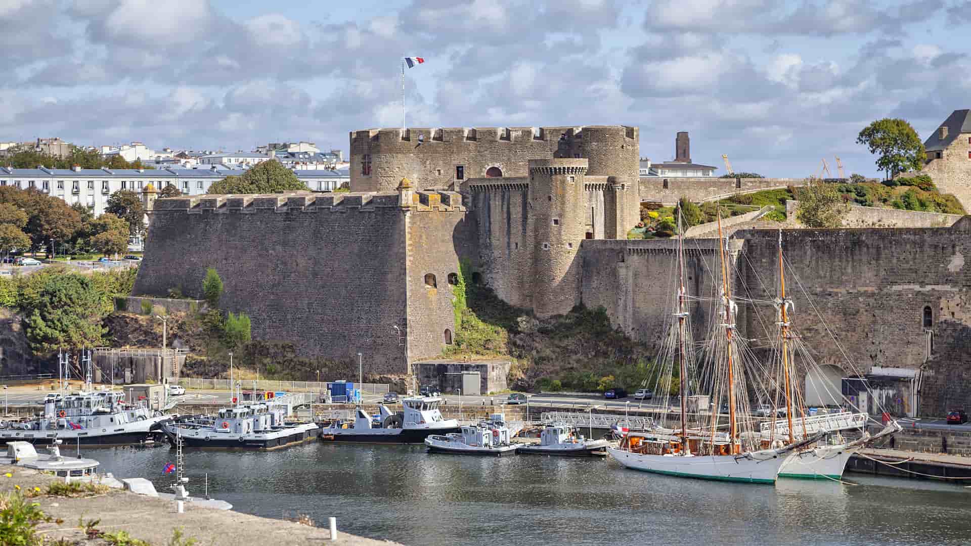 A beautiful port scene in Brest, France, with modern boats and a large sailboat docked in the foreground, and the historic medieval fortress, Château de Brest, towering over the harbor under a blue sky.