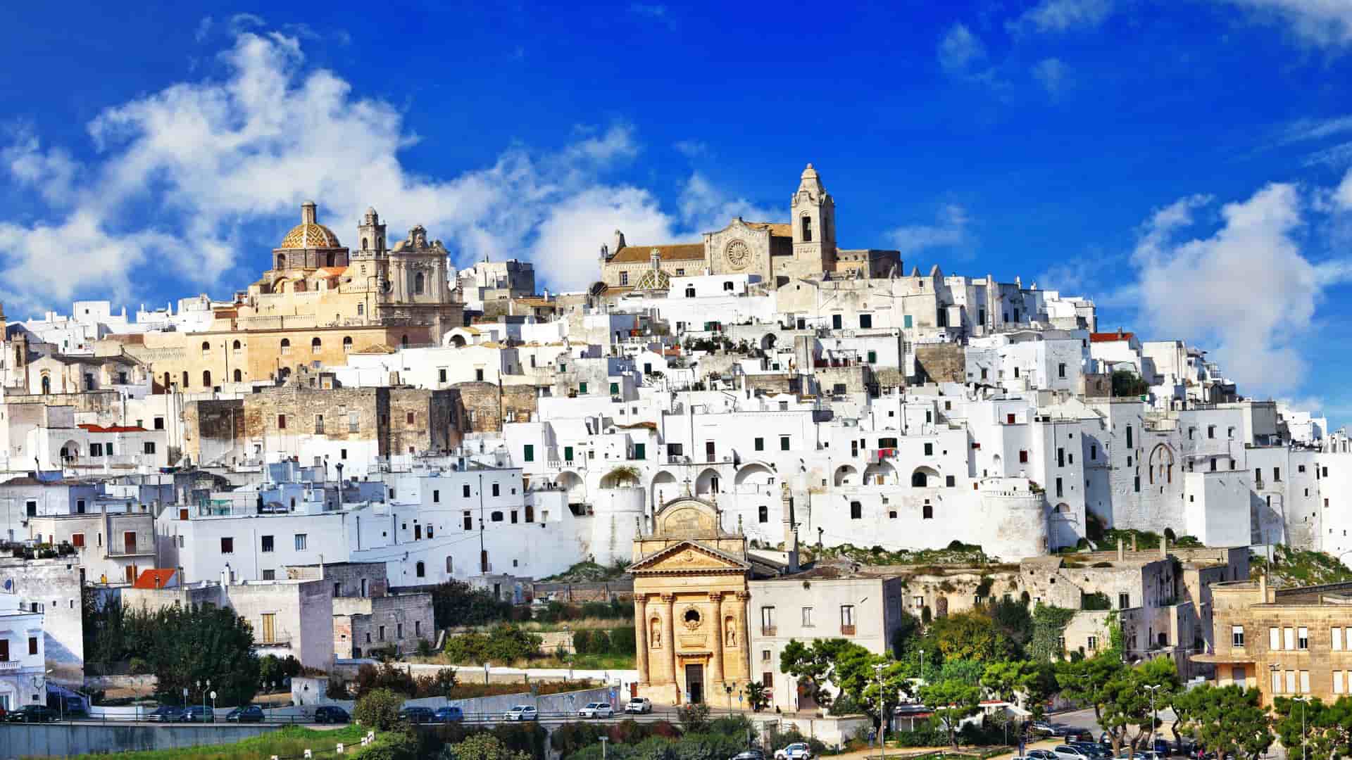 A vibrant panoramic shot of the beautiful white town of Ostuni, Italy, showcasing the historic buildings and churches on the hill under a brilliant blue sky with fluffy white clouds.