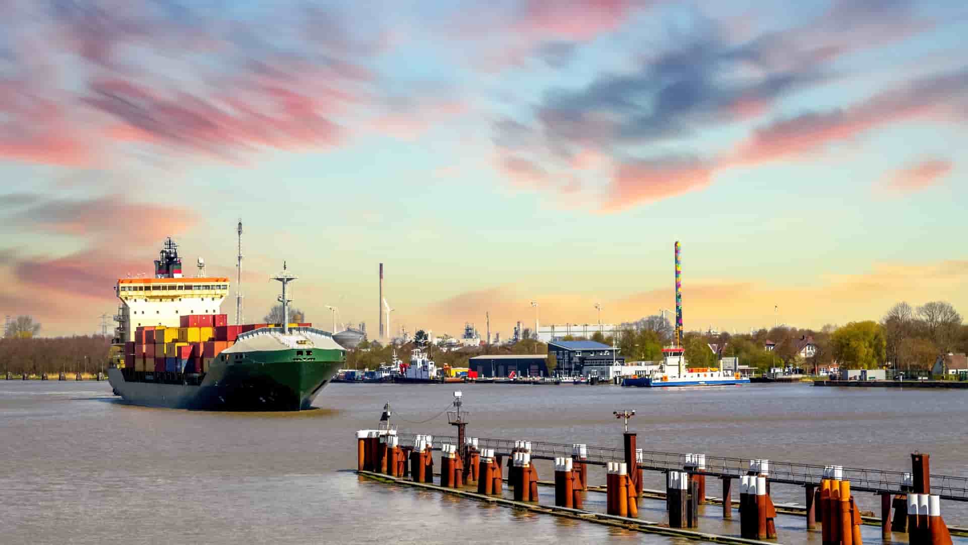 A large, green container ship moves along the water near a wooden dock in the harbor of Brunsbüttel, Germany, under a colorful sunset sky.