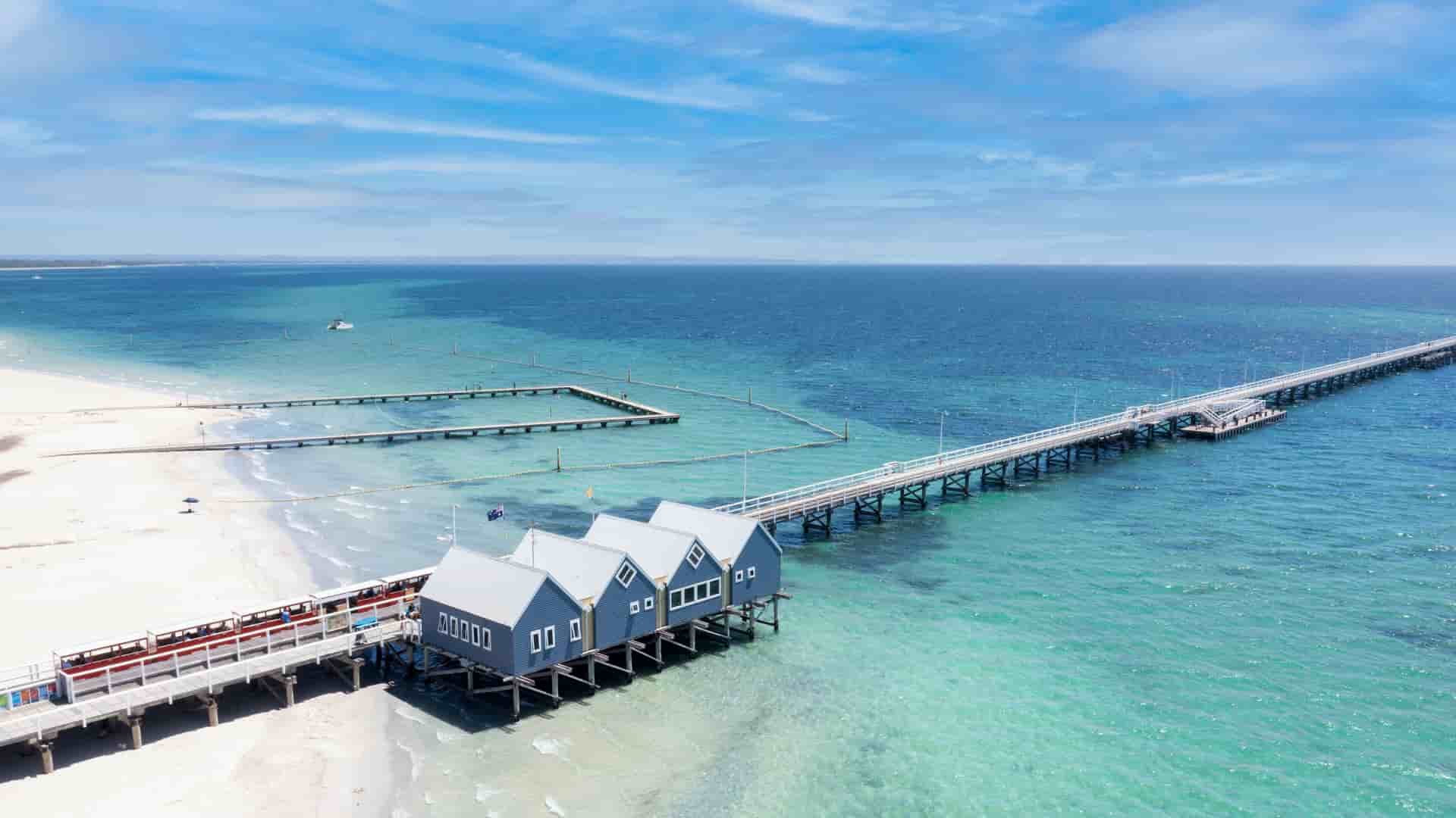 A scenic view of the iconic Busselton Jetty in Western Australia, with a row of small, blue-roofed buildings extending over the turquoise ocean.