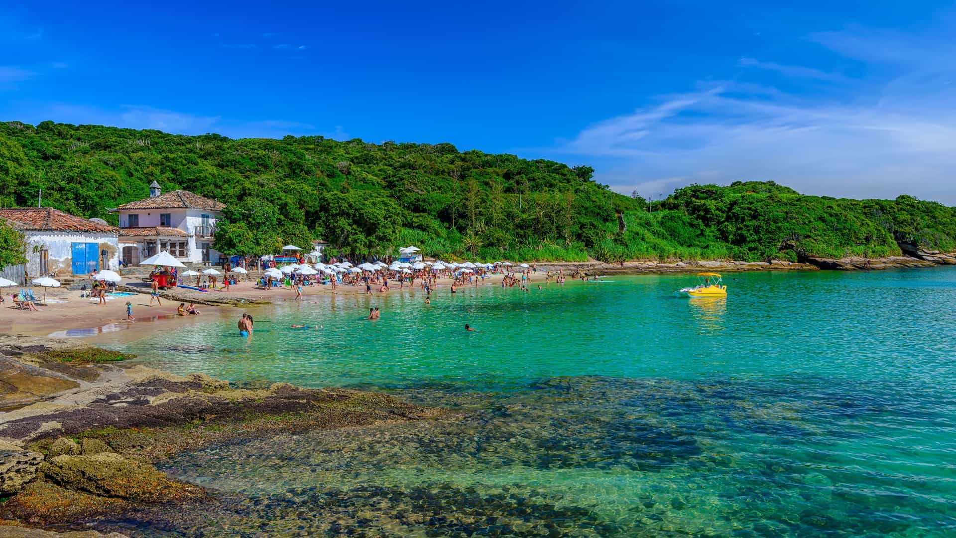 A vibrant beach scene in Búzios, Brazil, with people swimming and relaxing under white umbrellas on the sand, next to lush green hills and a small white building.