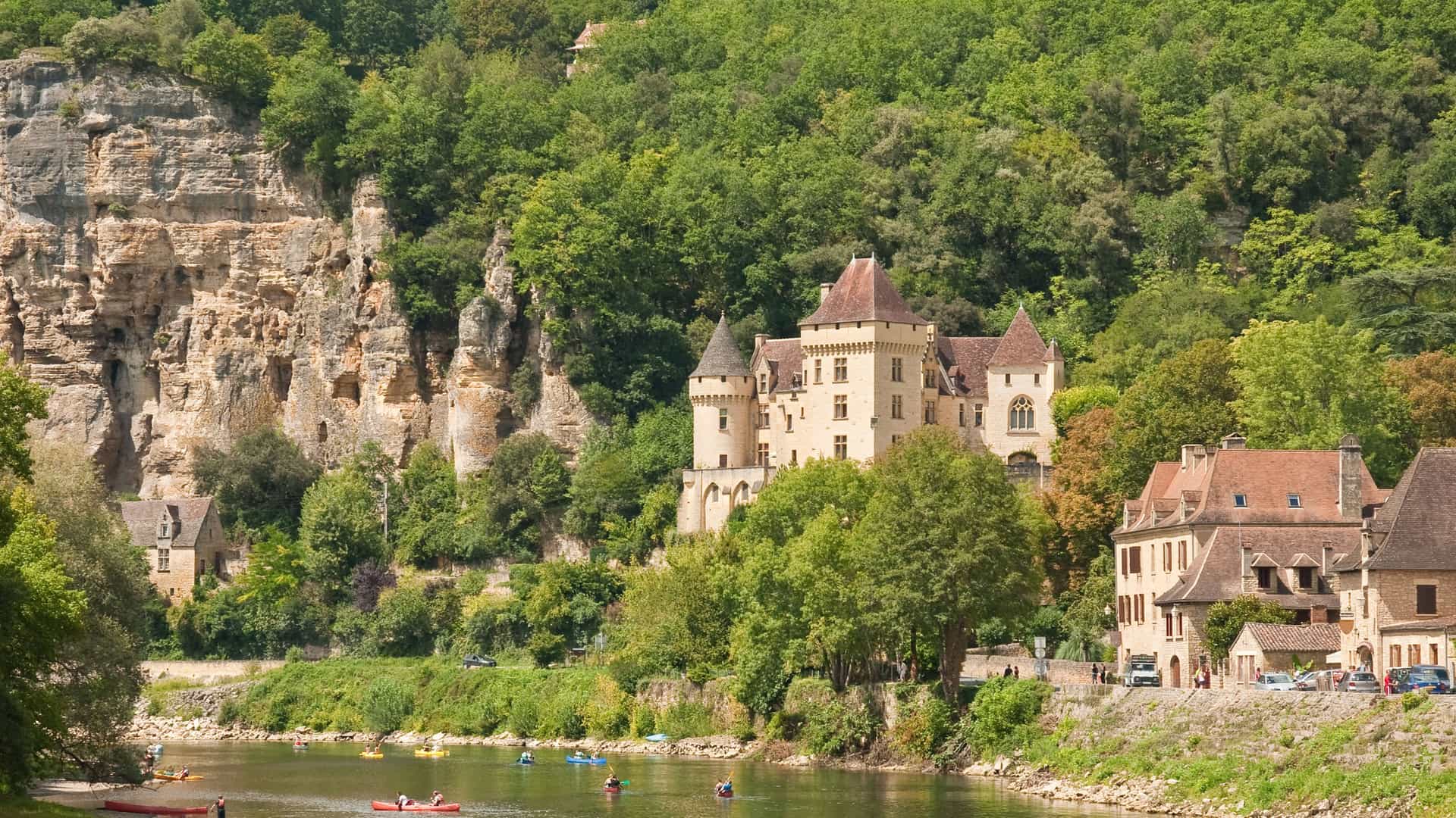 A scenic view of the Château de la Malartrie, a historic castle in Cadillac, France, nestled among the cliffs and lush trees beside the Dordogne River with people canoeing.