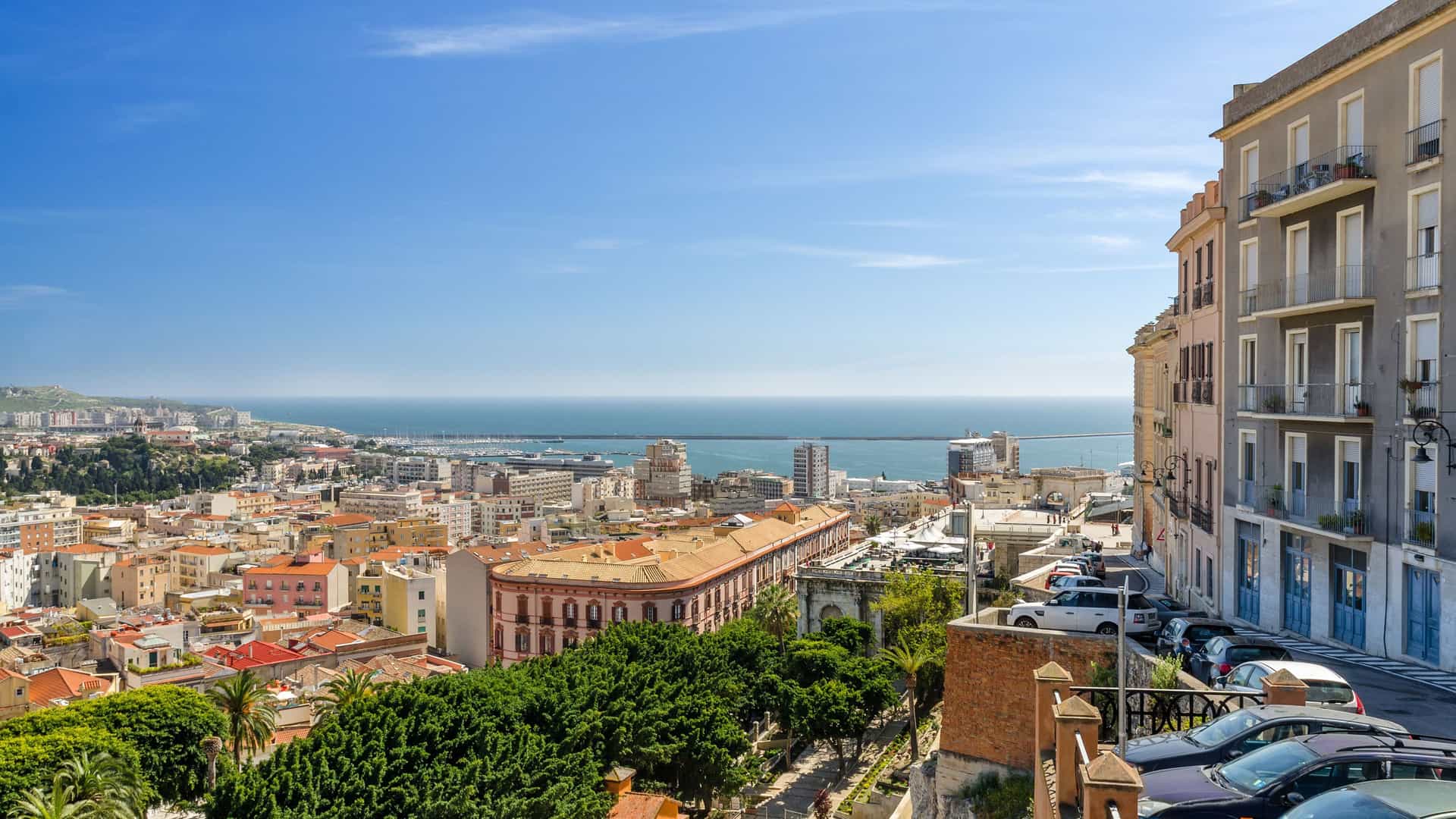 A high-angle view of the bustling city of Cagliari, Sardinia, Italy, with historic buildings and rooftops overlooking the Mediterranean Sea under a clear blue sky.