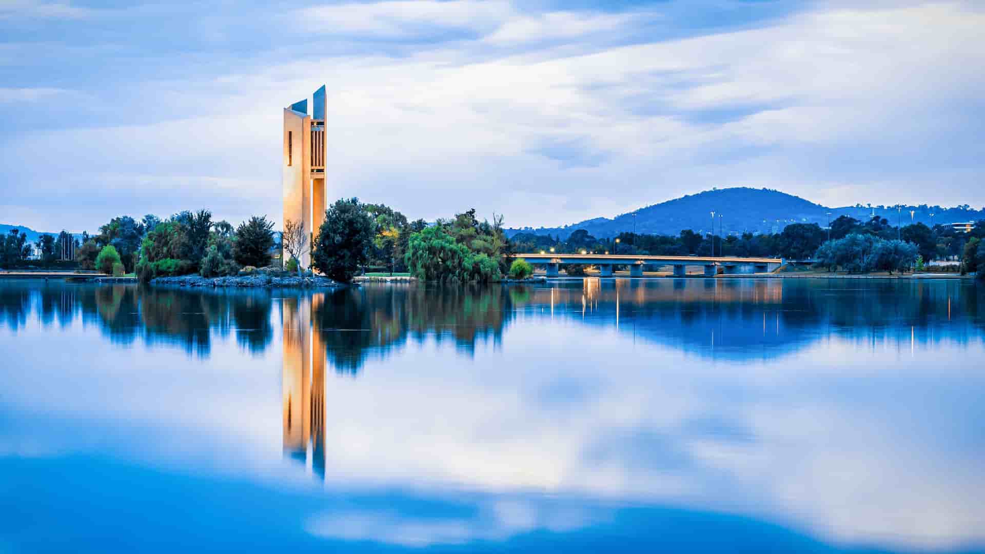 A serene, long-exposure photo of the National Carillon on Aspen Island in Lake Burley Griffin, Canberra, Australia, at dusk. The tall, modern bell tower is reflected perfectly in the smooth, glass-like surface of the lake, with a bridge and hills visible in the background.
