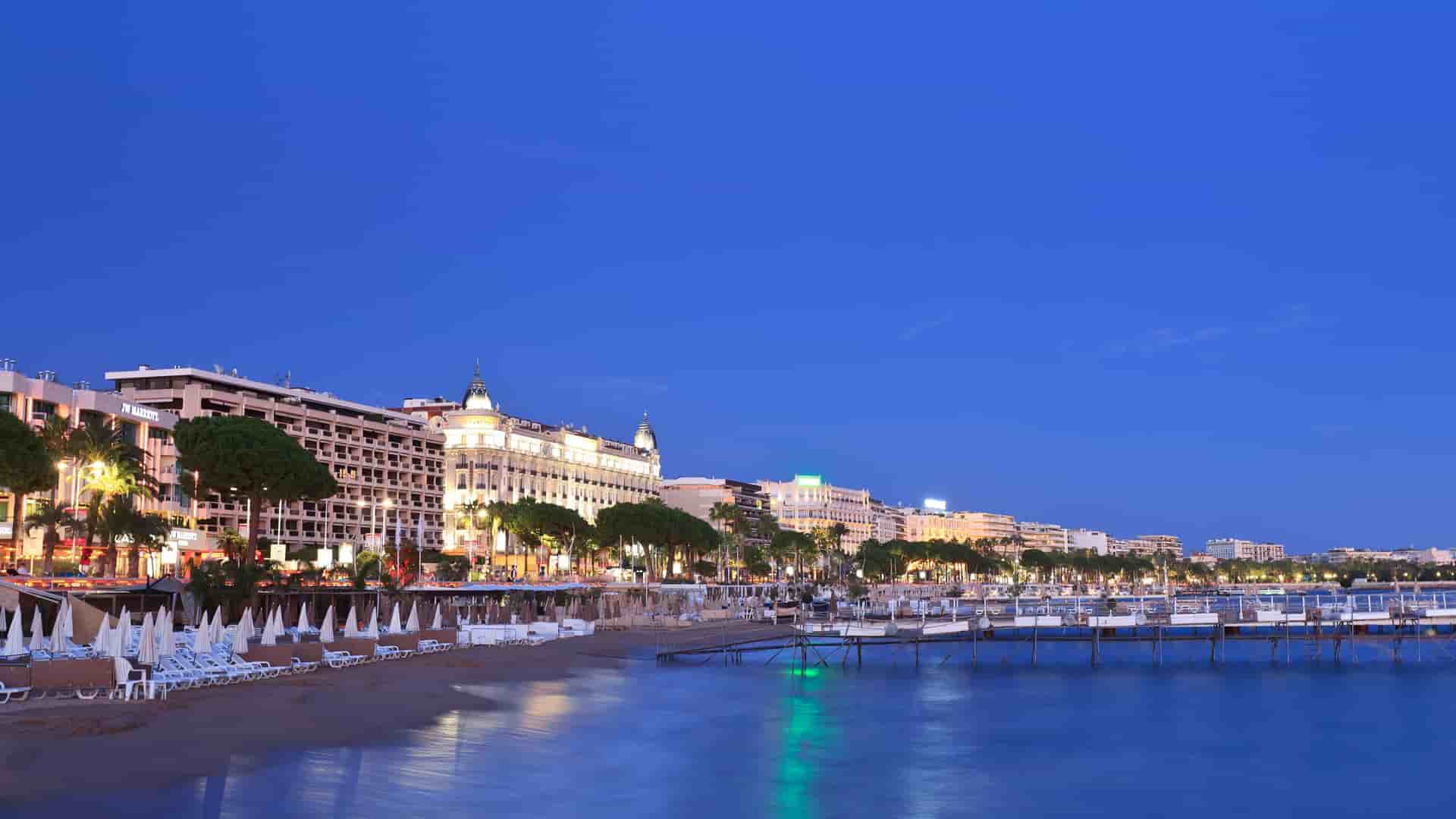 A wide-angle, twilight shot of the famous La Croisette promenade in Cannes, France, with its luxurious hotels, sandy beaches, and beautiful ocean views lit up at dusk.