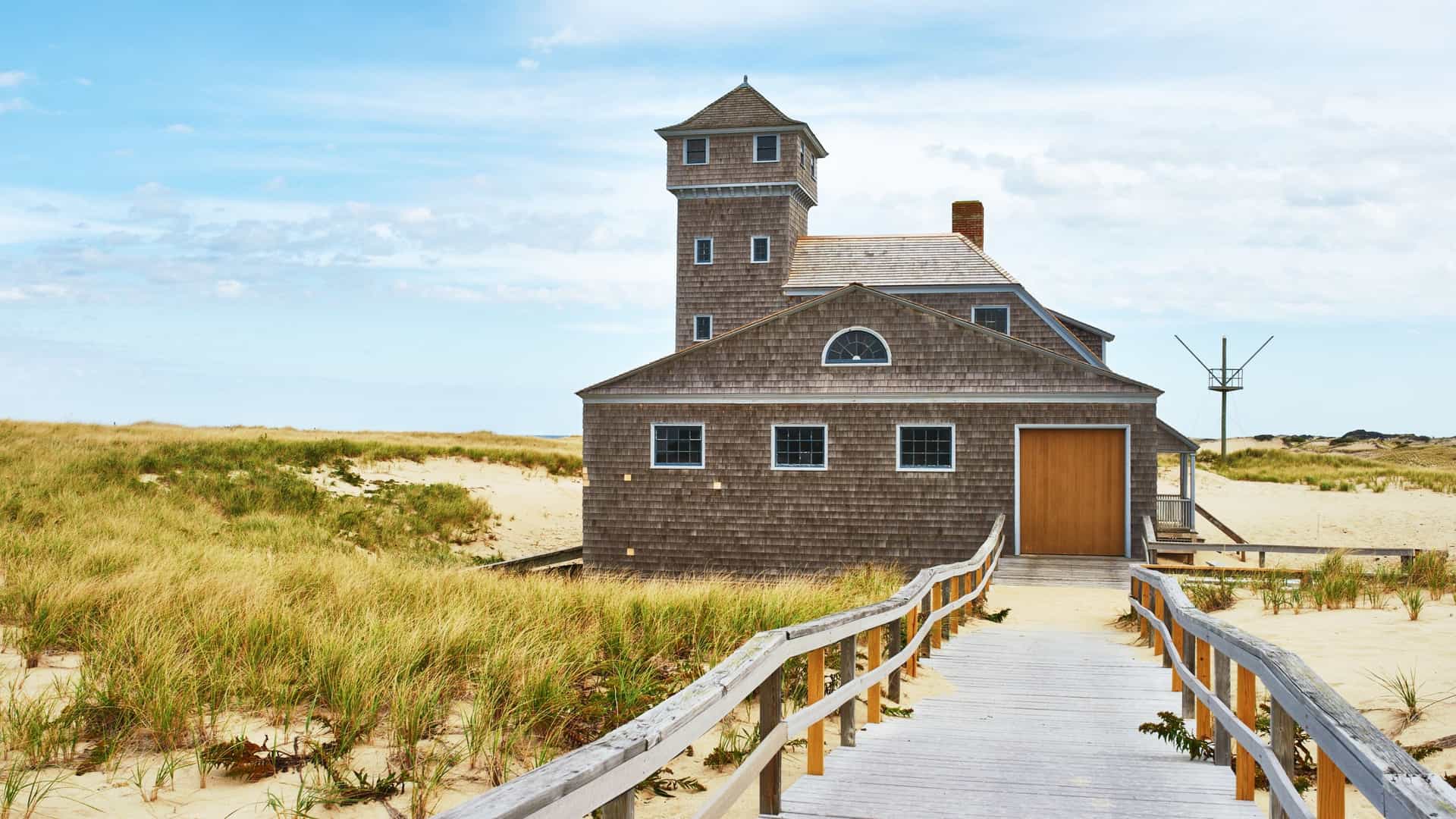 A shingled former life-saving station with a lookout tower sits in tall, dry grass and sand dunes on Cape Cod, Massachusetts, accessed by a wooden boardwalk.