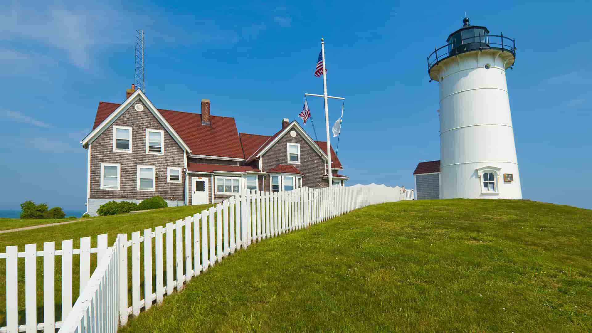 A beautiful daytime view of the iconic Nobska Lighthouse, a white lighthouse with a black top, and the attached keeper's house on a lush green hill with a white picket fence on Cape Cod, Massachusetts.