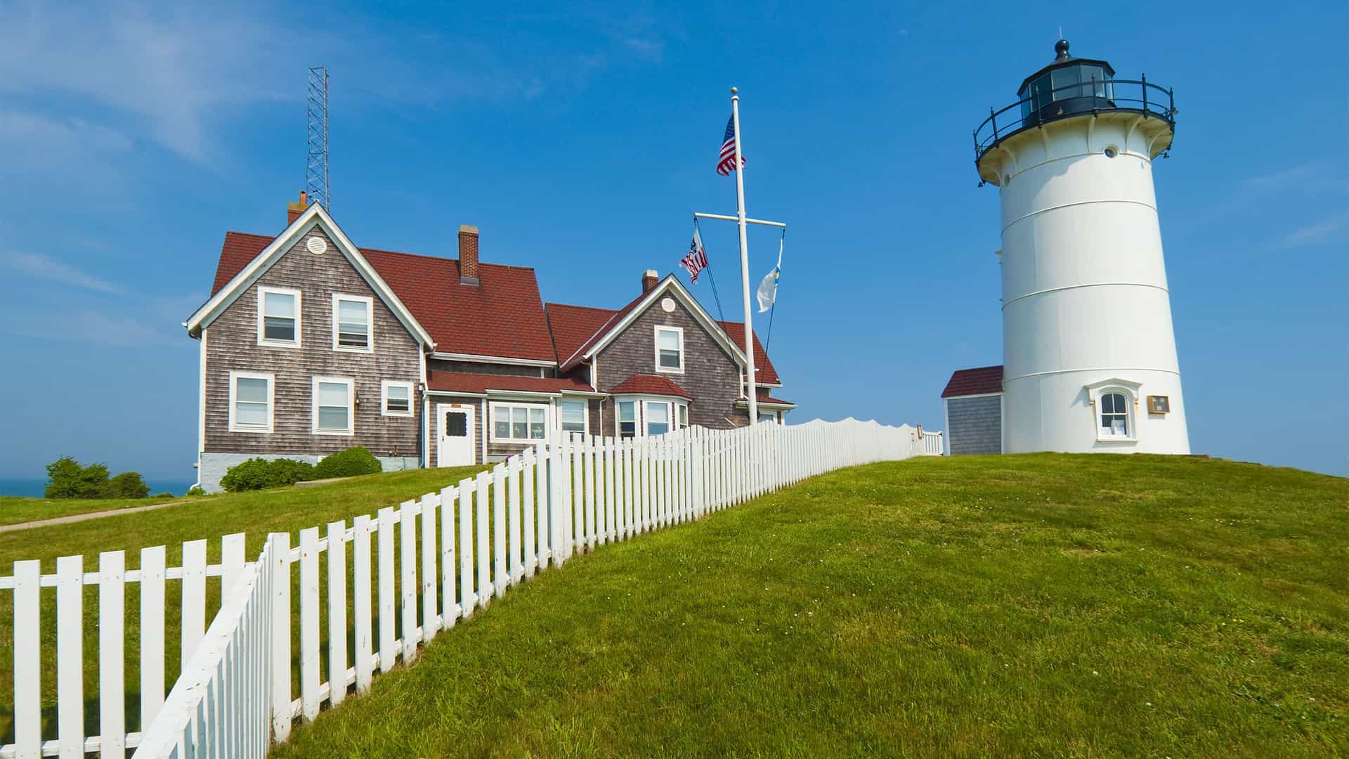 A beautiful daytime view of the iconic Nobska Lighthouse, a white lighthouse with a black top, and the attached keeper's house on a lush green hill with a white picket fence on Cape Cod, Massachusetts.