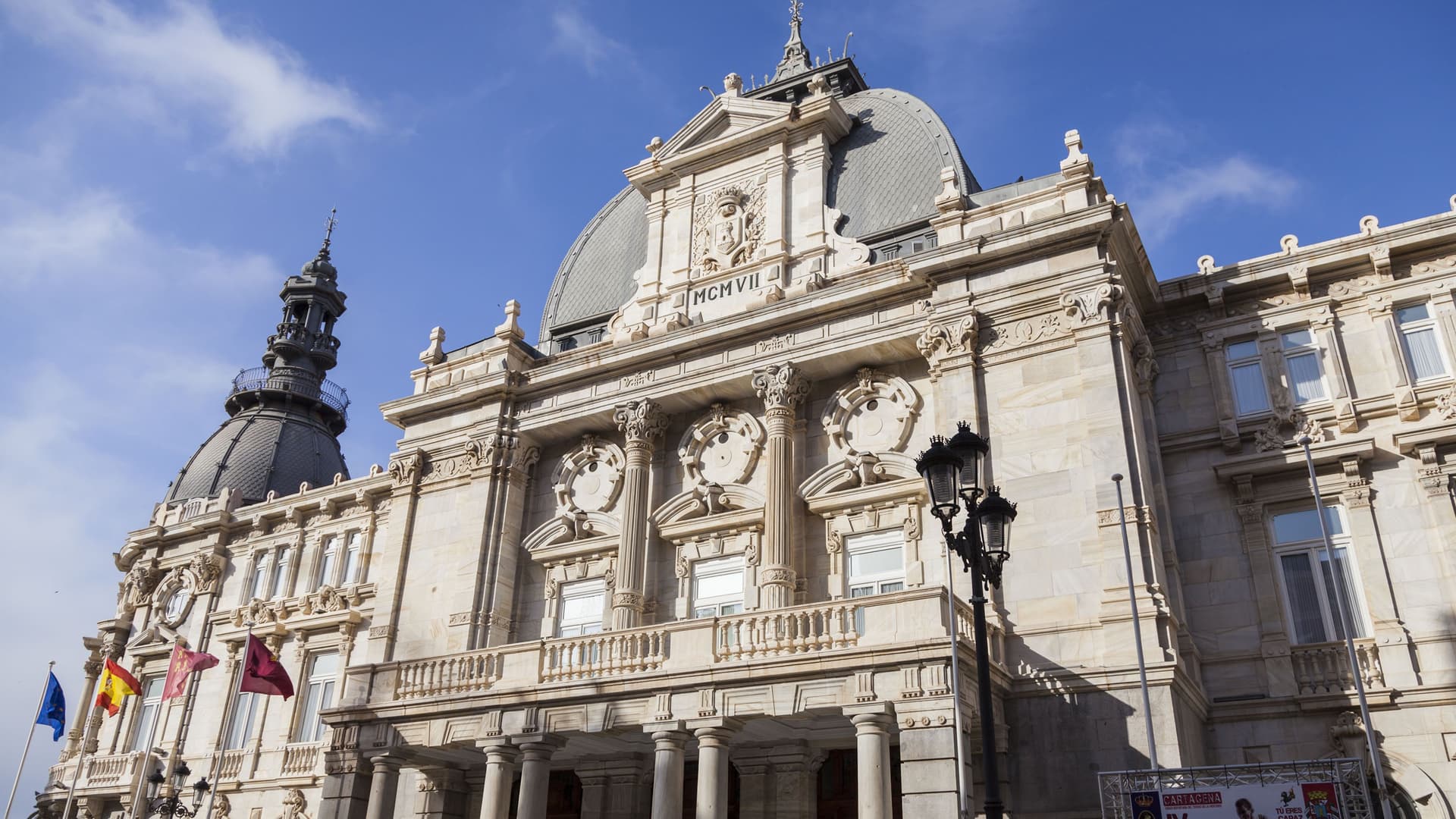 A low-angle shot of the magnificent Palacio Consistorial, the town hall of Cartagena, Spain, showcasing its ornate, white marble facade and domed rooftops against a blue sky.
