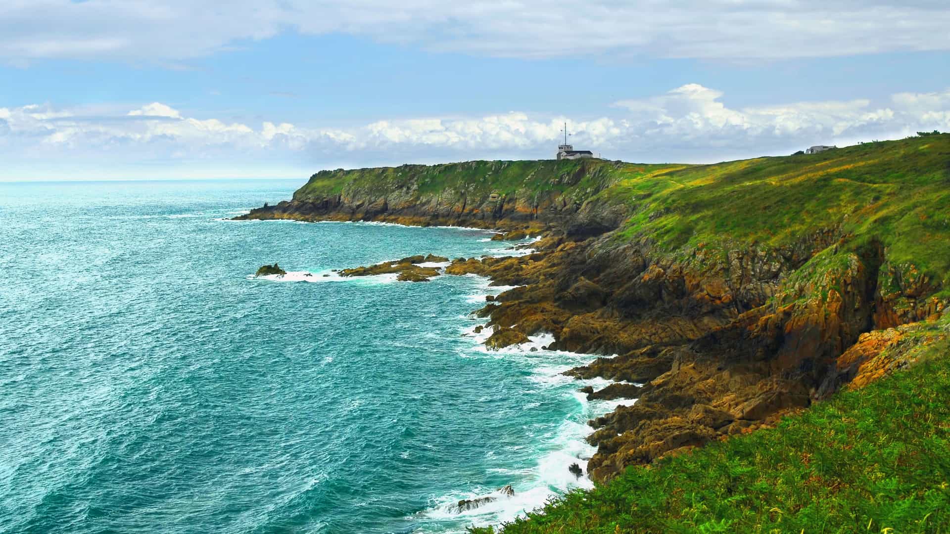 A rugged, green cliffside overlooking the choppy turquoise waters of the sea, with a small white lighthouse visible on the horizon.