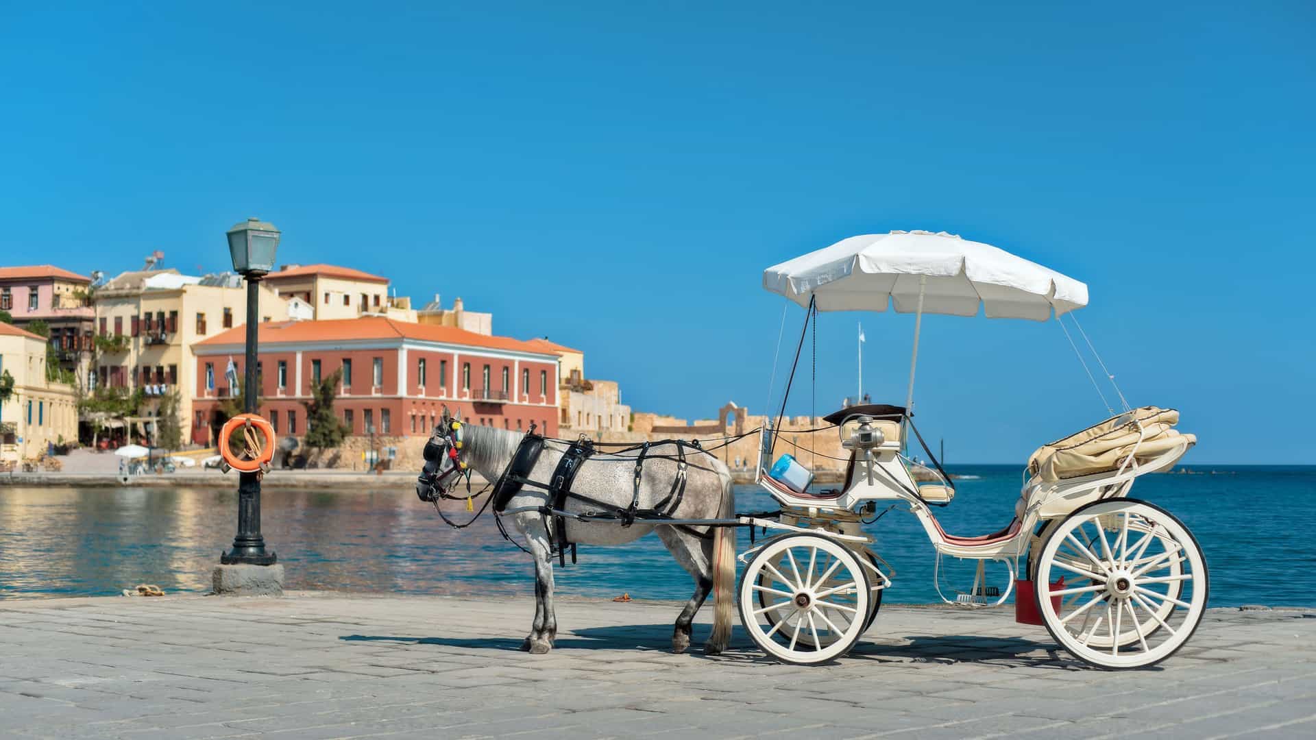 A picturesque scene of a horse-drawn carriage on the waterfront of Chania, Crete, with historic Venetian harbor buildings and the deep blue Mediterranean Sea in the background.