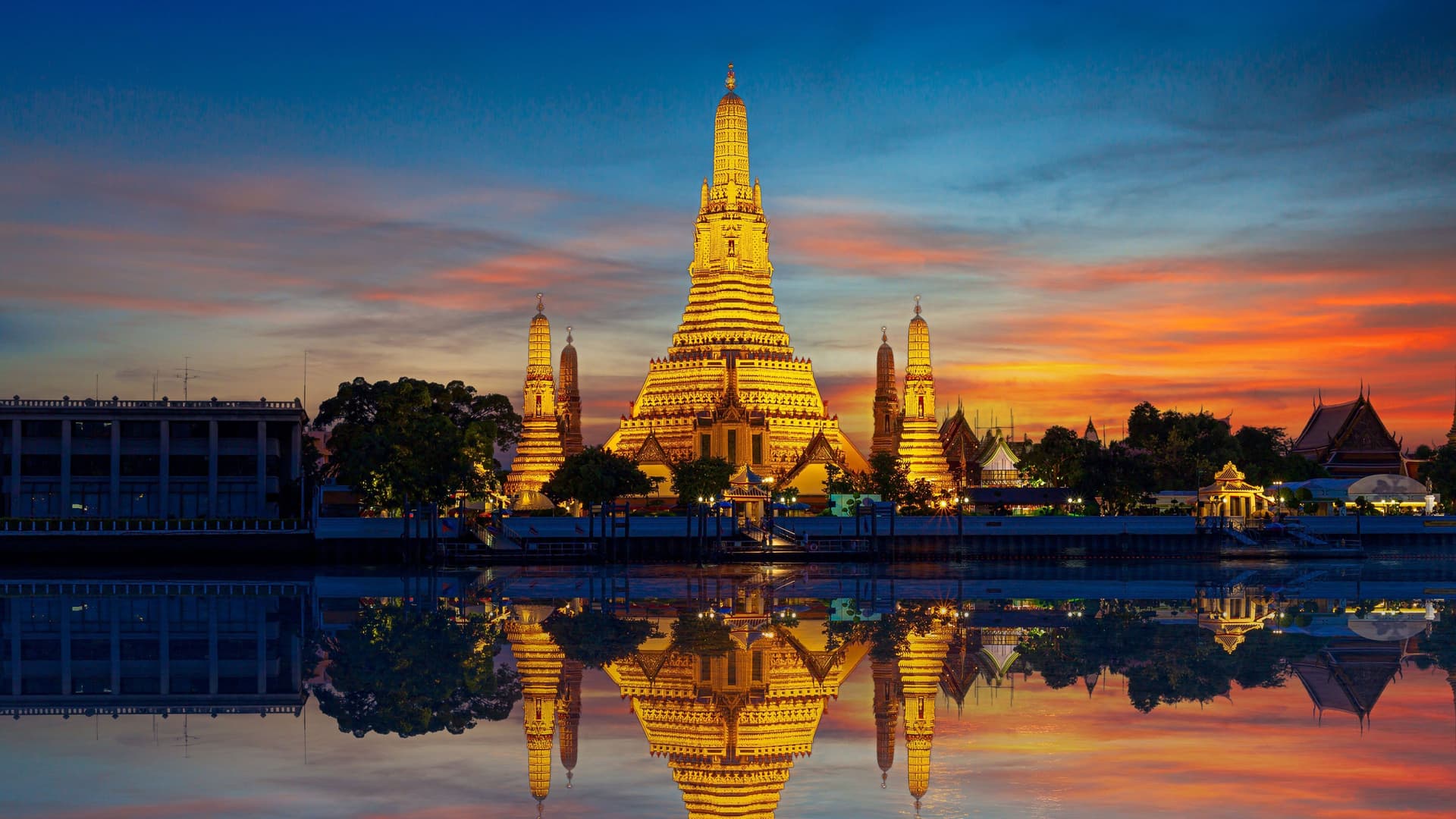 A vibrant sunset view of the illuminated Wat Arun temple, known as the Temple of Dawn, reflecting on the surface of the Chao Phraya River in Bangkok, Thailand.