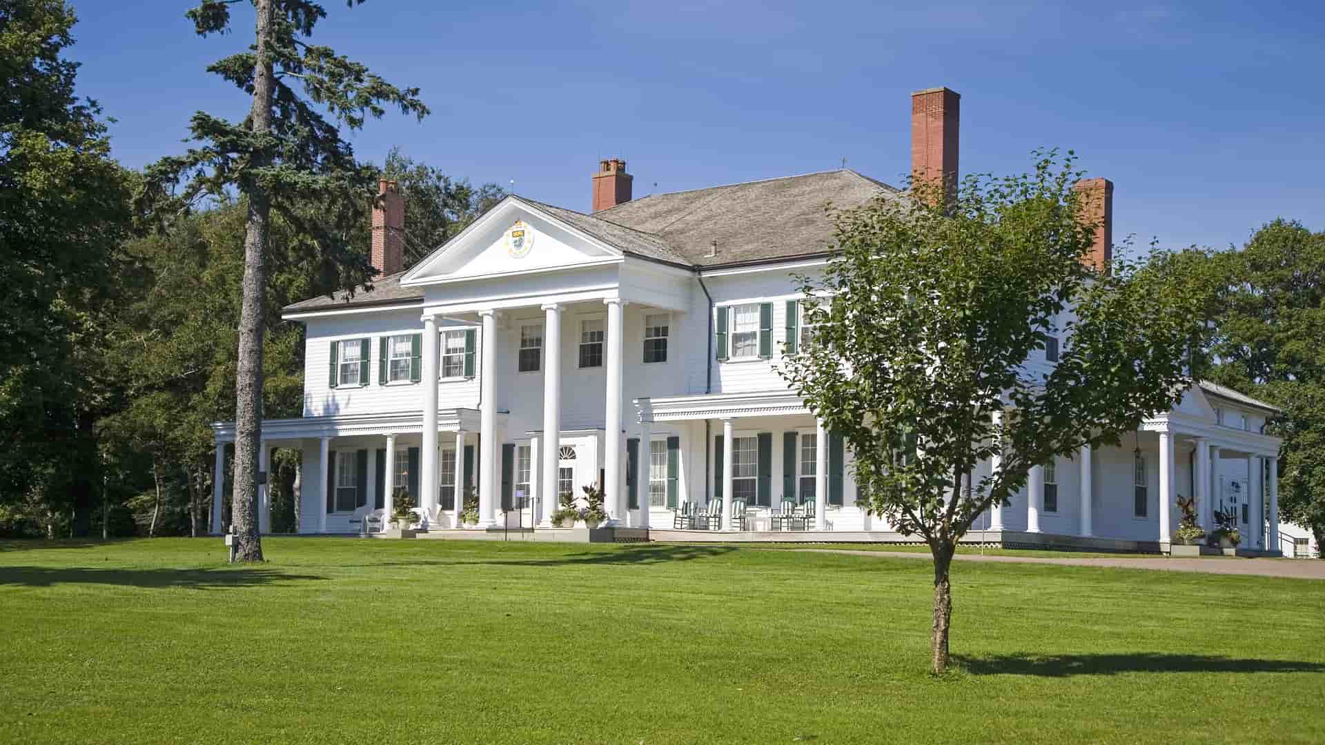 A wide shot of the Government House, a large white mansion with a Greek Revival facade and a prominent portico, surrounded by a green lawn and tall trees in Charlottetown.
