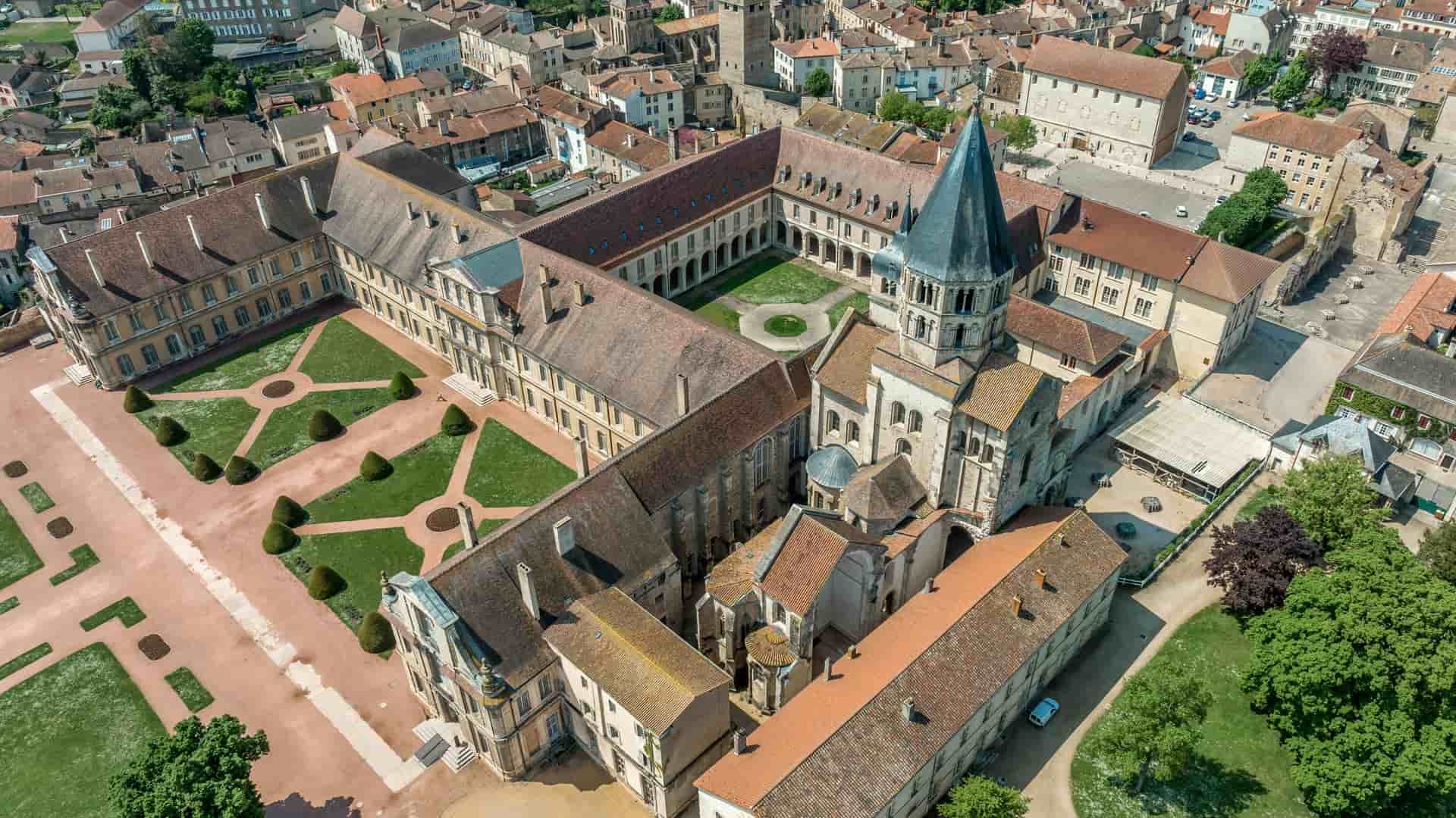 An aerial view shows the extensive architectural complex of Cluny Abbey, France, with the abbey ruins and surrounding buildings in a dense townscape.